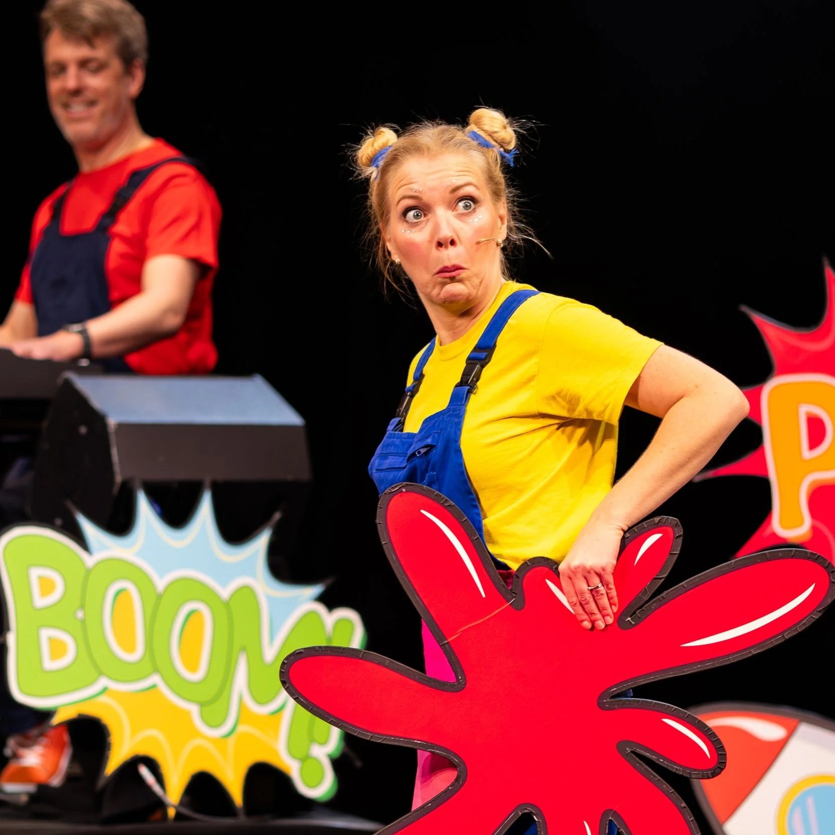 Two performers on stage, one woman in a yellow shirt with blue overalls and her hair in two buns, making a surprised expression, and one man in a red shirt with black overalls, smiling behind colorful comic-style signs that say 'BOOM!'.