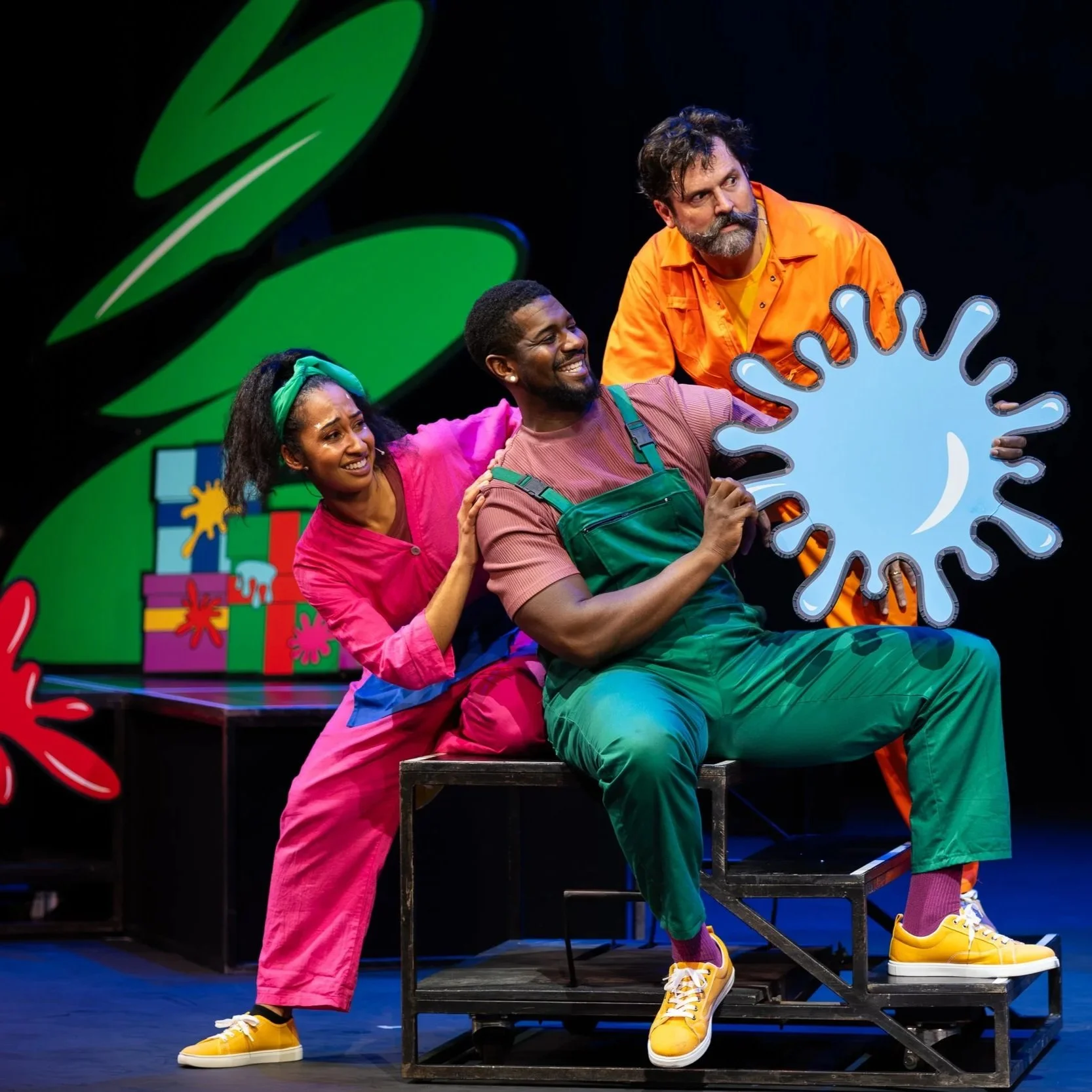 Three actors perform on stage, with colorful costumes and stage props including a large green abstract shape and painted boxes resembling gifts. One actor holds a large blue splash cutout, and they are all expressing playful emotions.