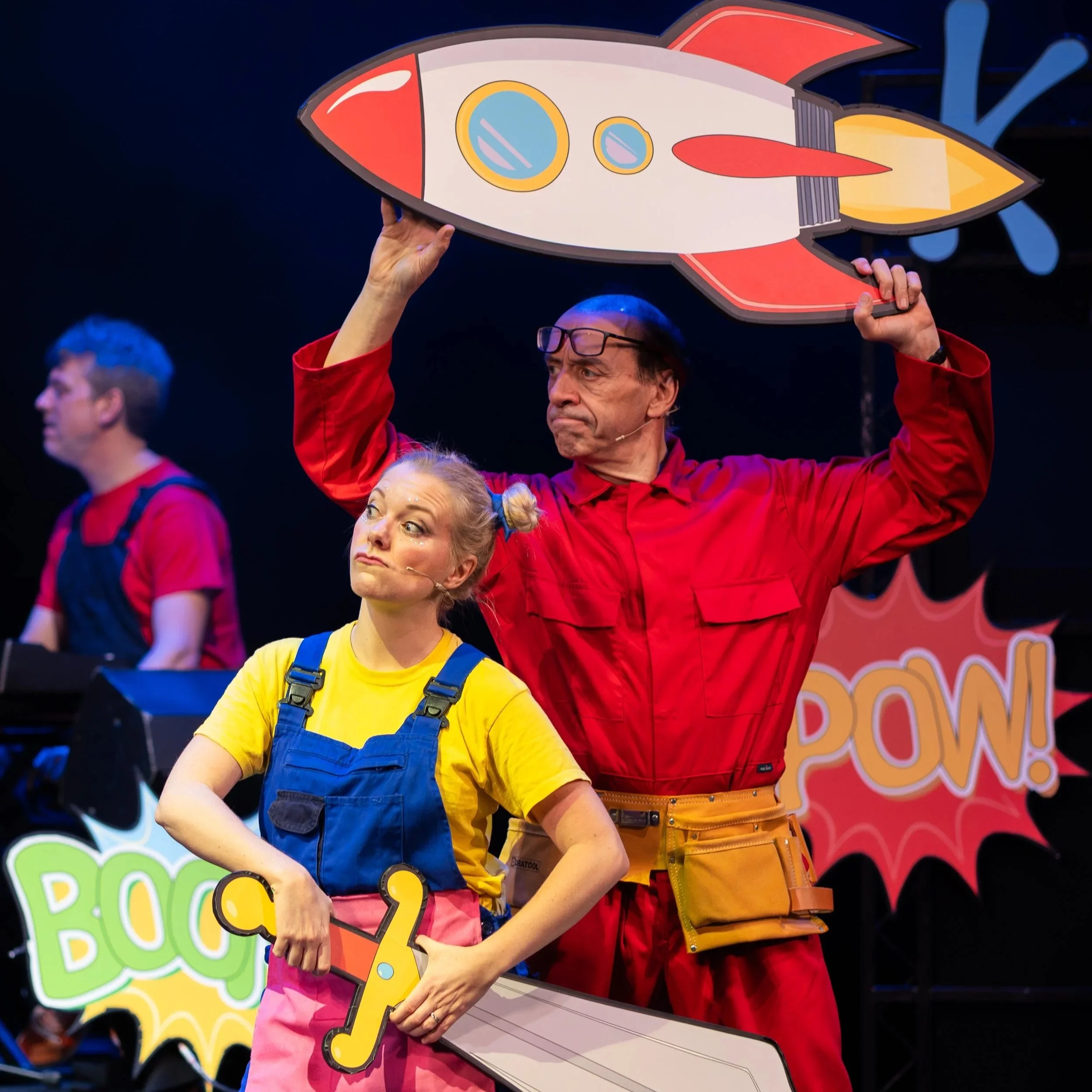 Three performers on stage dressed as superheroes, with comic book style props and speech bubbles reading 'BOOM!' and a large rocket cutout.