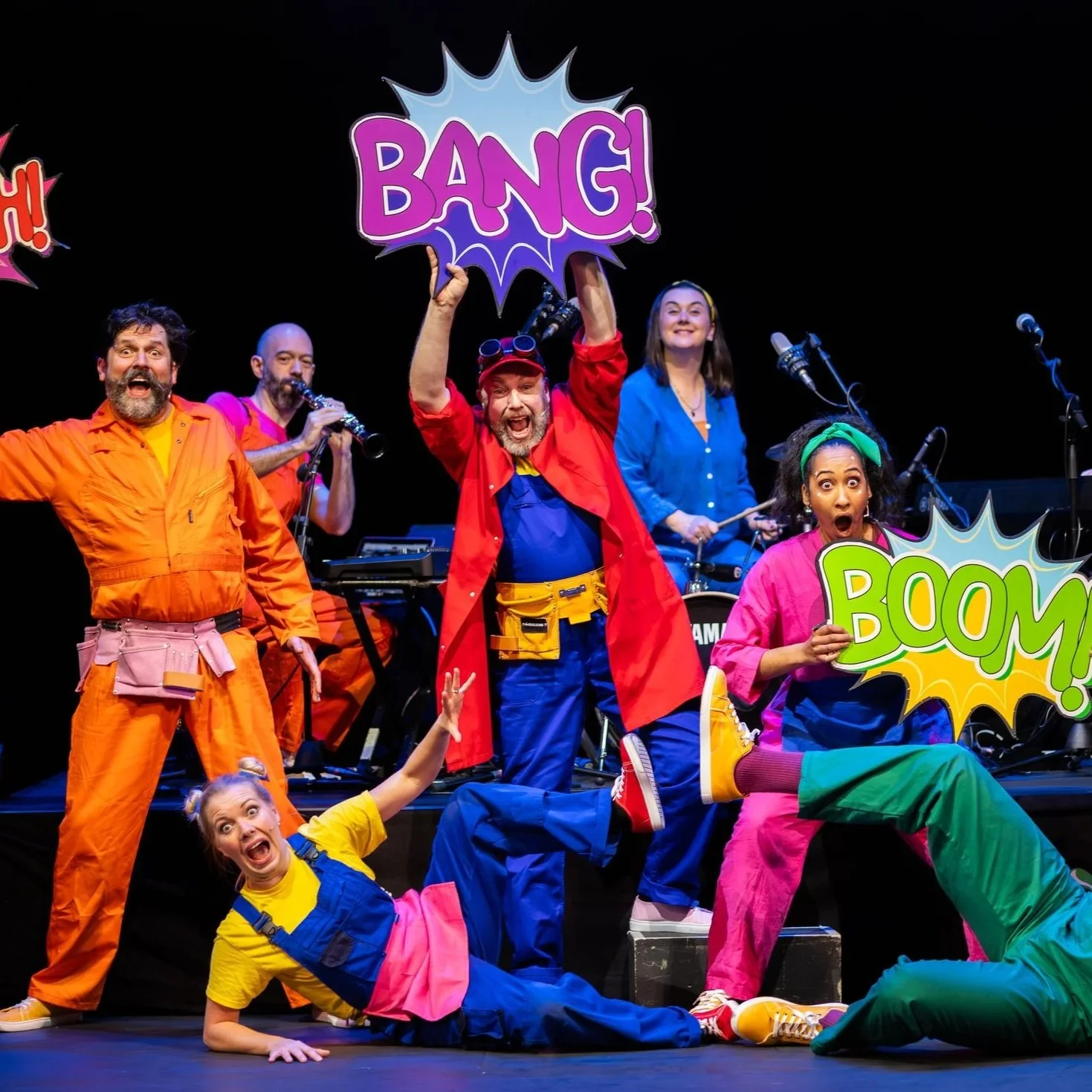 Group of actors dressed in colorful costumes performing on stage, holding comic-style signs with words like 'BANG!', 'BOOM!', and 'BANG!'. They are posing energetically, with some lying on the stage and others standing, creating a lively, playful sce