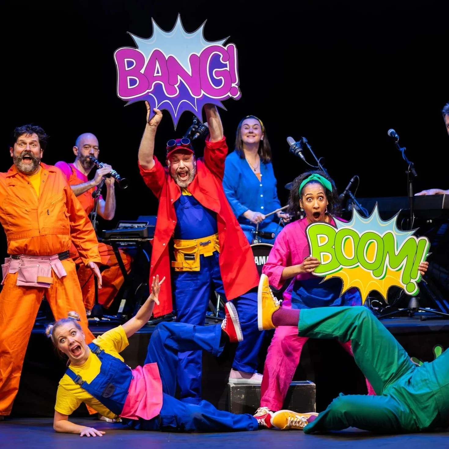 A lively stage performance featuring a diverse cast of actors in colorful costumes, holding comic book style signs saying 'BANG!', 'BOOM!', and others, with musical instruments in the background, expressing excitement and surprise.