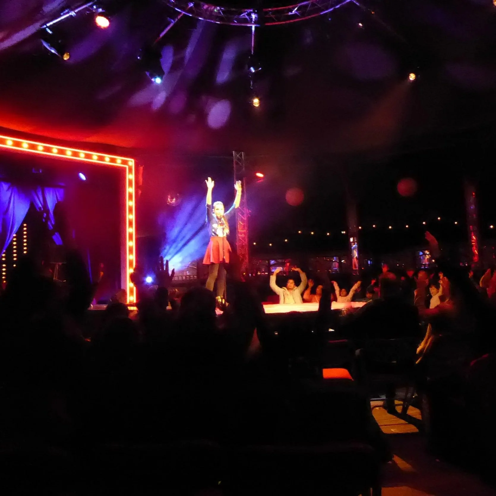 A lively stage performance with a woman standing on stage, raising her arms, surrounded by audience members with hands raised. The stage is illuminated with colorful lights, and there are several performers seated in the background.