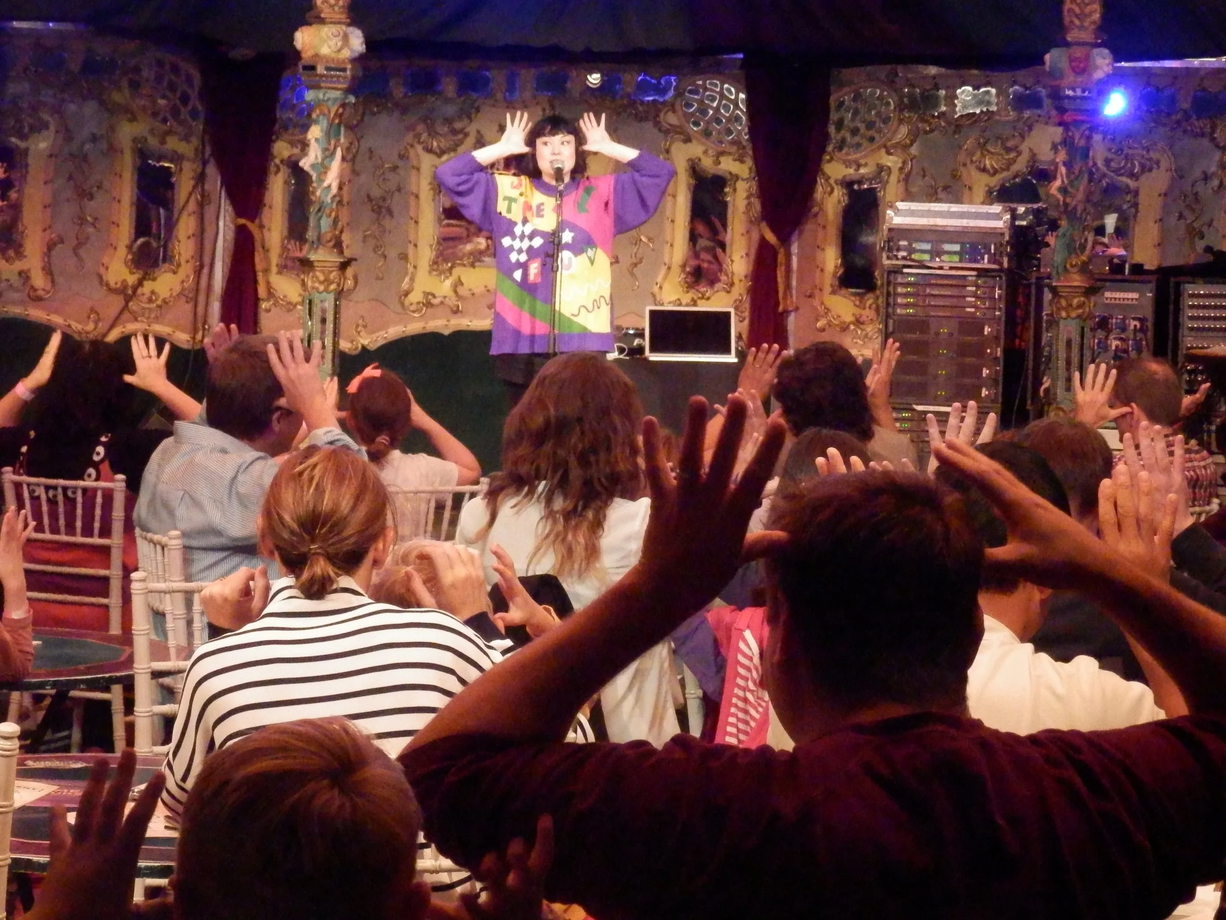 A performer on stage with colorful, patterned clothing and a microphone, engaging with an audience who have their hands raised, in an ornate, decorated venue.