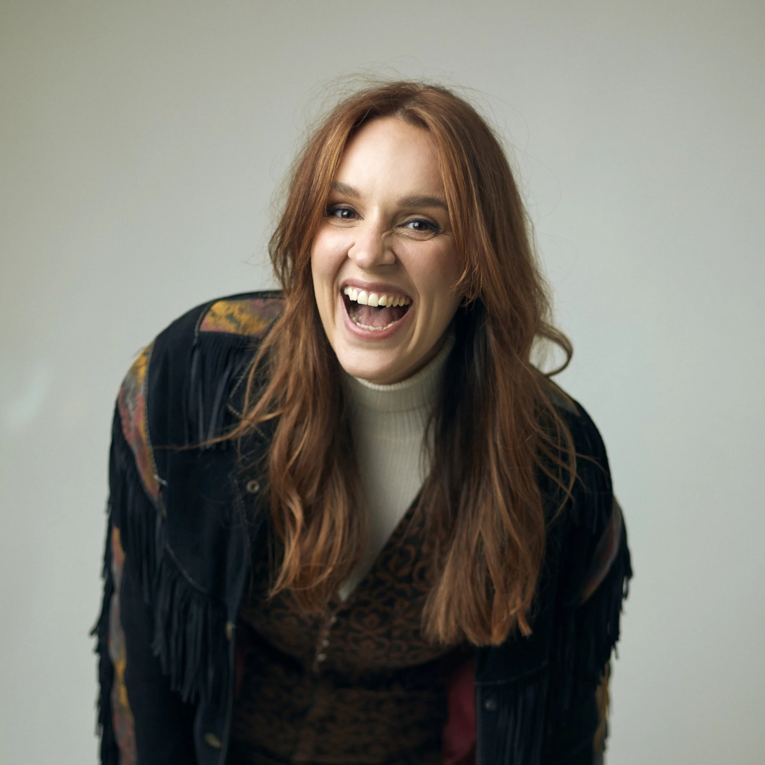 A woman with long red hair singing or shouting with a joyful expression, wearing a black jacket with a floral pattern on the shoulder, standing against a plain light gray background.