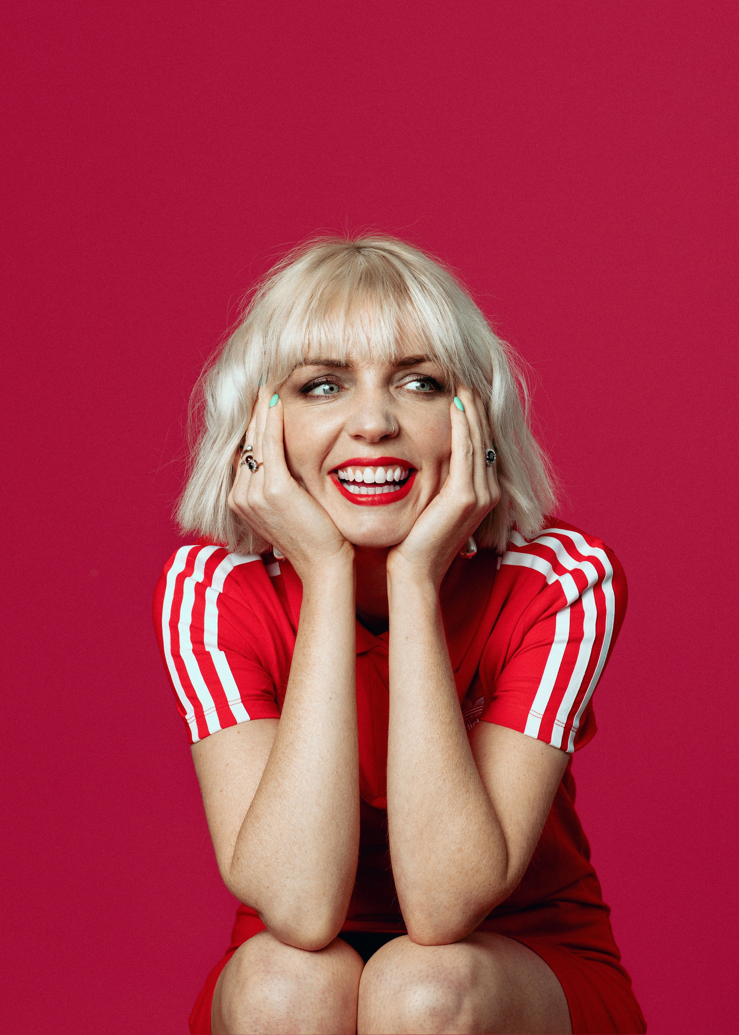 A woman with blonde hair, red lipstick, and a nose ring, wearing a red sports jersey with white stripes, sitting against a red background, smiling and resting her face on her hands.