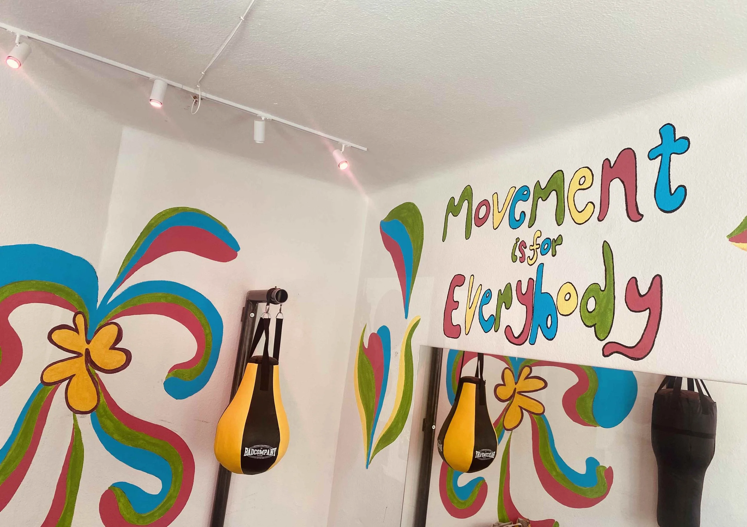Colorful mural on wall with painted flowers, swirls, and the words 'movement is for everybody' in rainbow colors, with boxing gloves hanging from the ceiling.