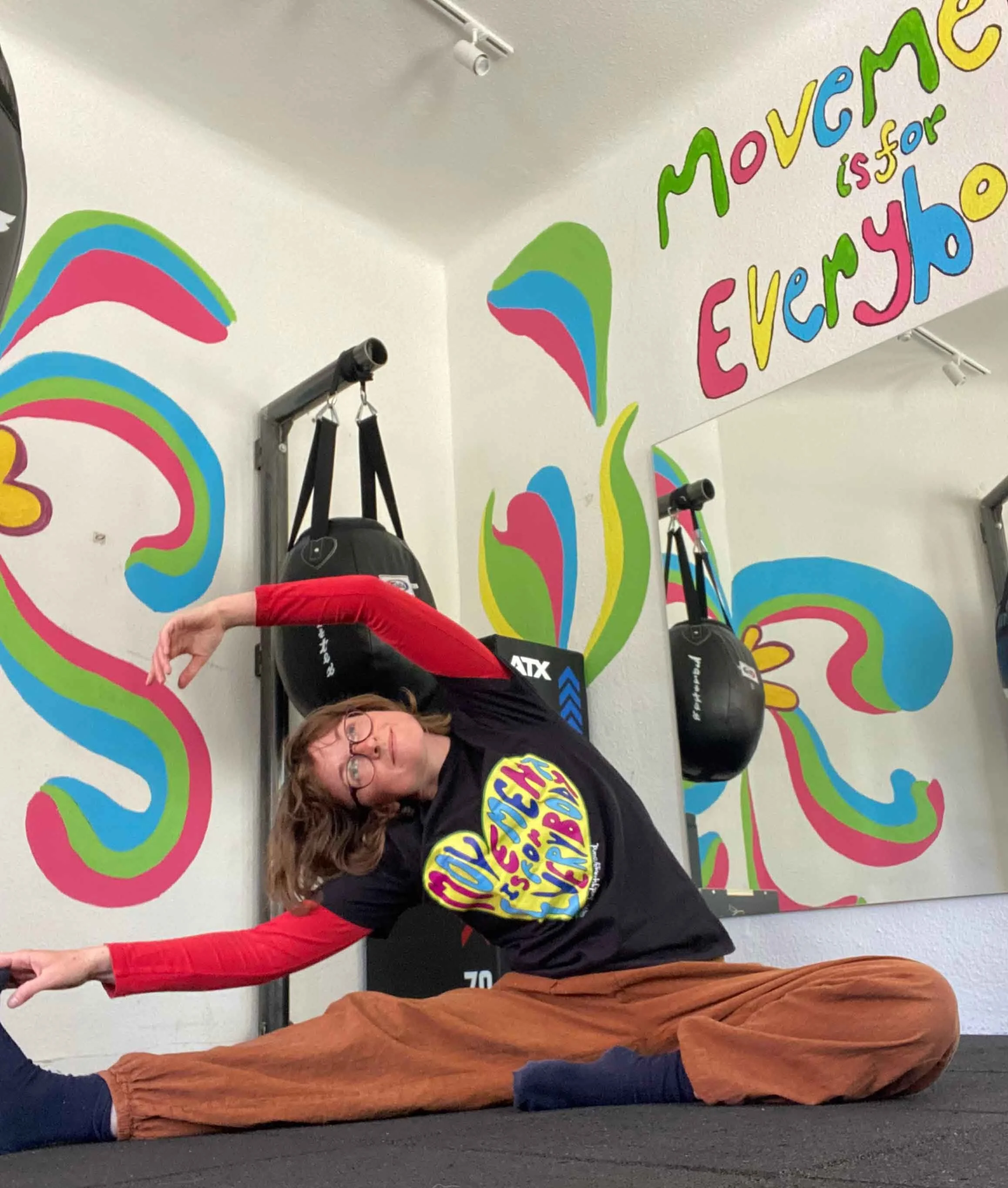 A person performing a stretching exercise in a fitness studio with colorful wall art and motivational words, wearing workout clothes and glasses.