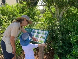 Adult and child reading an outdoor informational sign surrounded by trees and greenery.