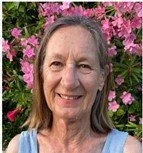 A woman outdoors with pink flowering bushes in the background, smiling at the camera.