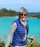Older woman in sunglasses, blue vest, and gray sweater standing by a turquoise sea with trees in the background.