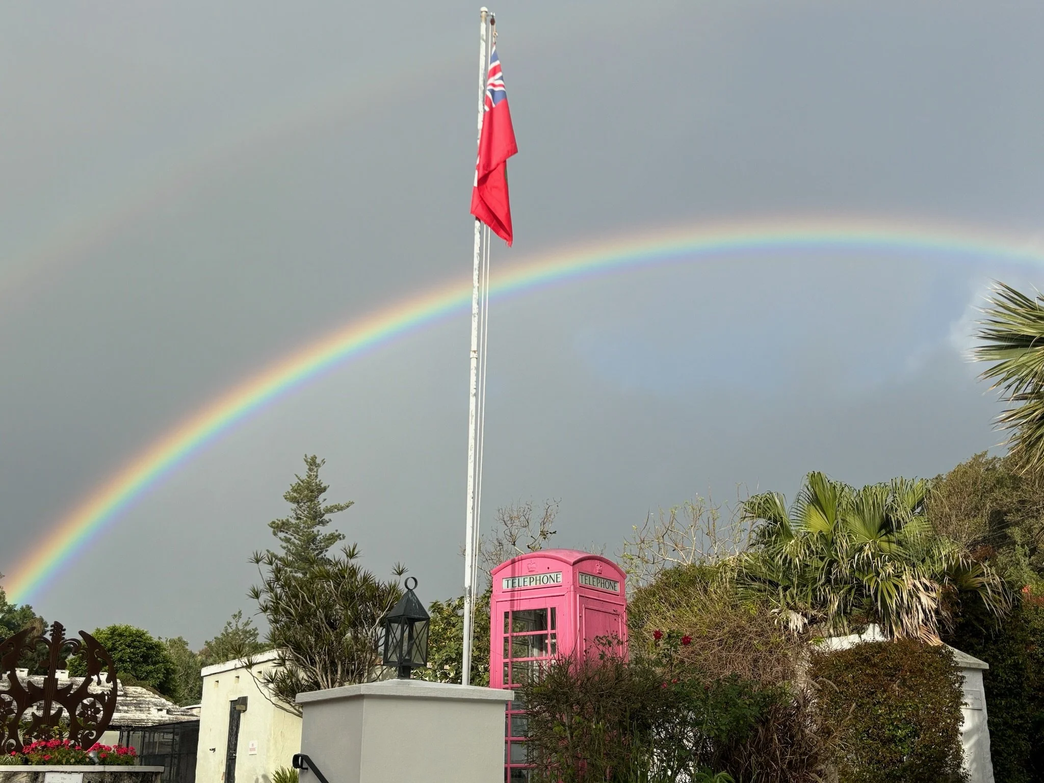 A double rainbow in the sky with a cloudy background. In the foreground, there is a bright pink vintage telephone booth, a flagpole with a red flag, and various green trees and plants.