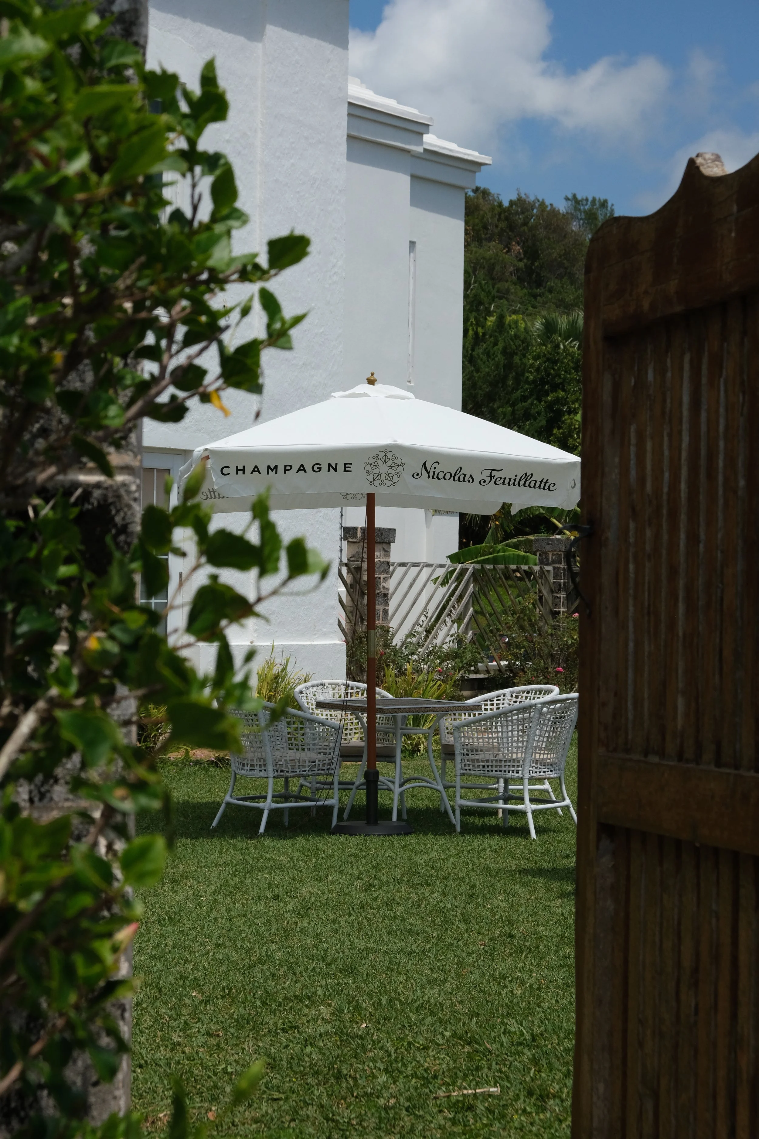 A backyard patio with a round table, four white woven chairs, and a large white umbrella that says 'Champagne Nicolas Feuillatte'. The patio is partly obscured by a wooden gate and some green bushes, with a white house and blue sky in the background.