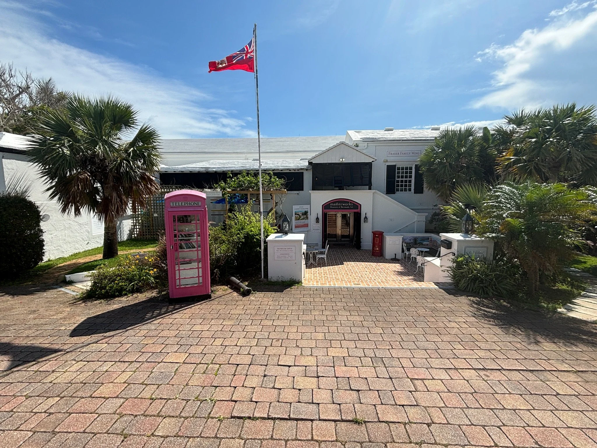 A white building with black shutters, a staircase, and outdoor seating, surrounded by palm trees and plants, with a pink telephone booth in the foreground, a red mailbox, and a flag flying on a pole under blue sky with some clouds.