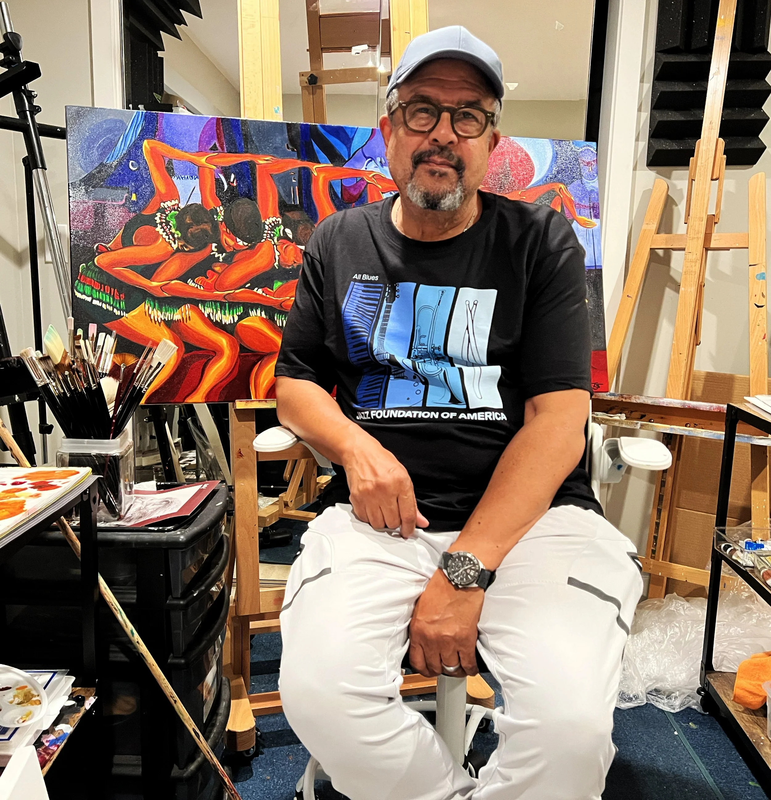 A man wearing a black T-shirt with a jazz-themed graphic, beige pants, and glasses sitting on a white stool in an art studio, with a colorful painting behind him depicting figures dancing.