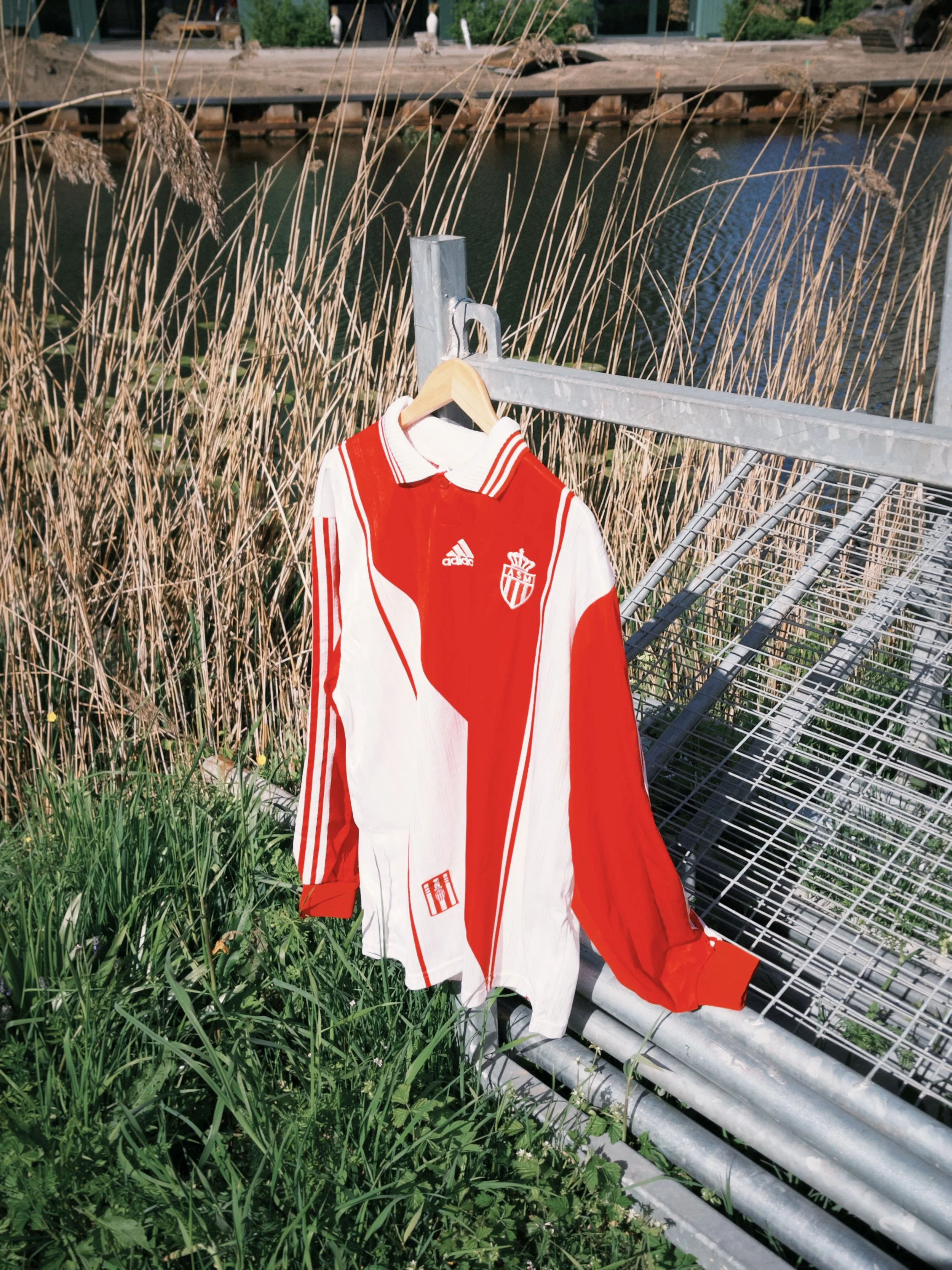 A soccer jersey with red and white colors hanging on a hanger on a metal railing near green grass and plants, with a body of water and some buildings in the background.