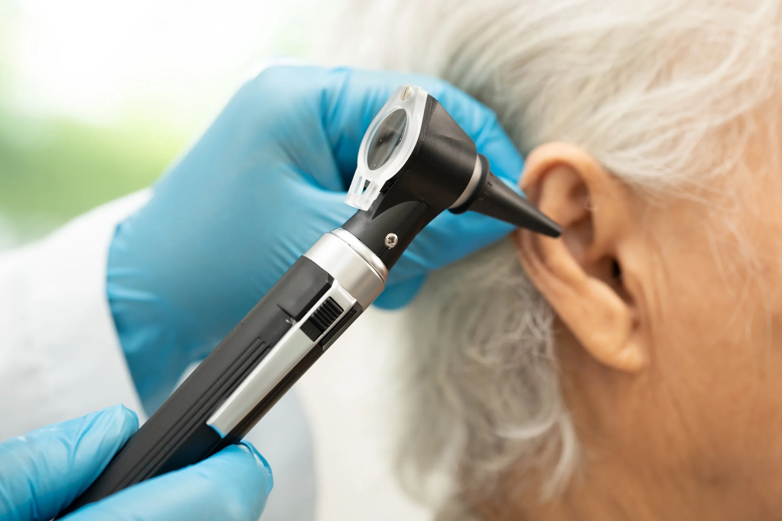 A healthcare worker wearing blue gloves uses an otoscope to examine an elderly person's ear.
