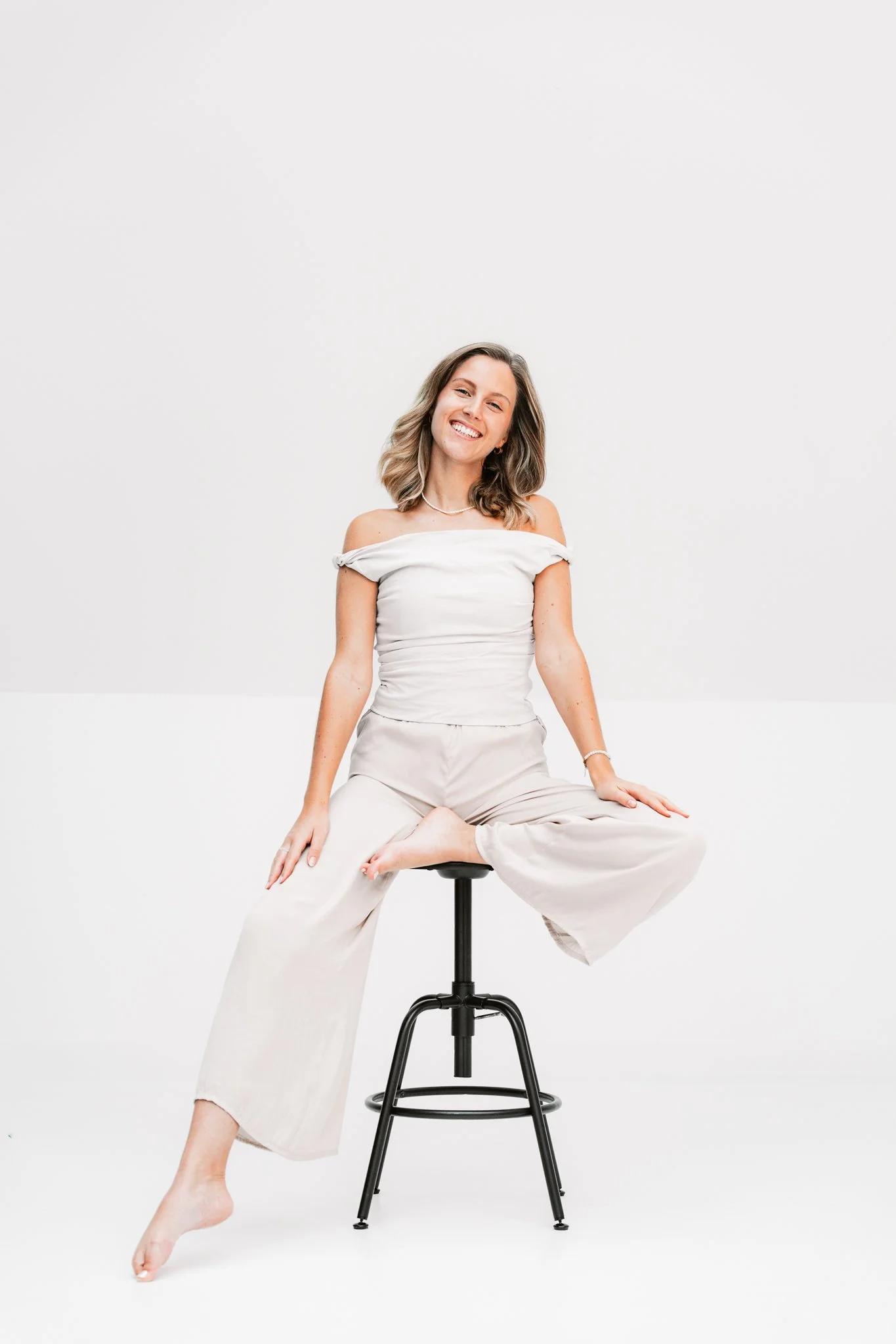 Junge Frau sitzt auf einem Hocker, trägt weiße Kleidung, lächelt und wirkt entspannt.