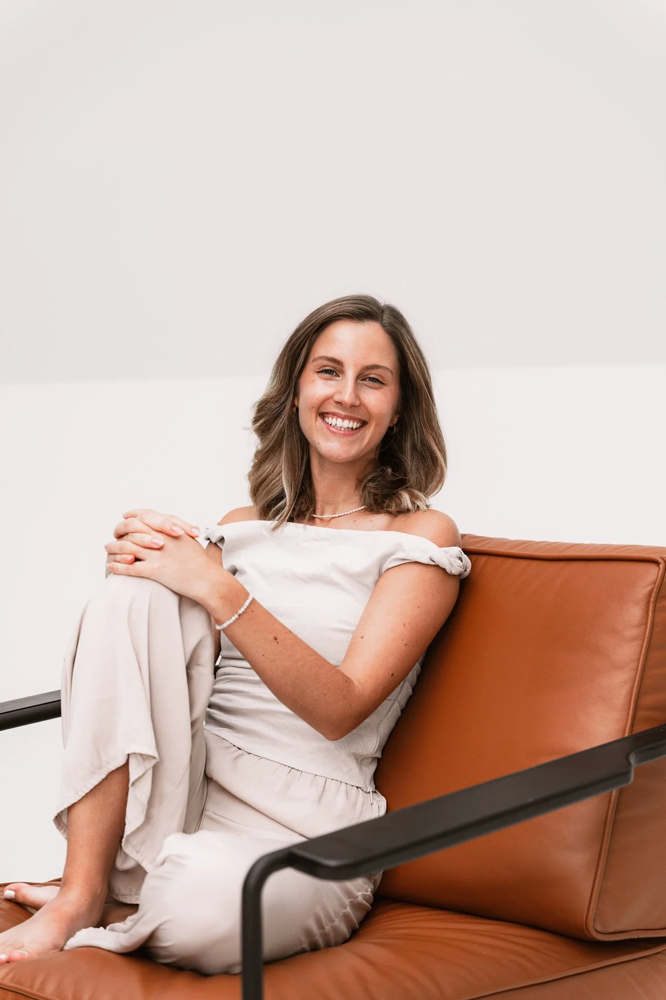 Eine lachende junge Frau sitzt gemütlich auf einem braunen Sofa vor einer weißen Wand, trägt ein weißes off-shoulder T-Shirt, Schmuck und hat braune Wellenhaare.