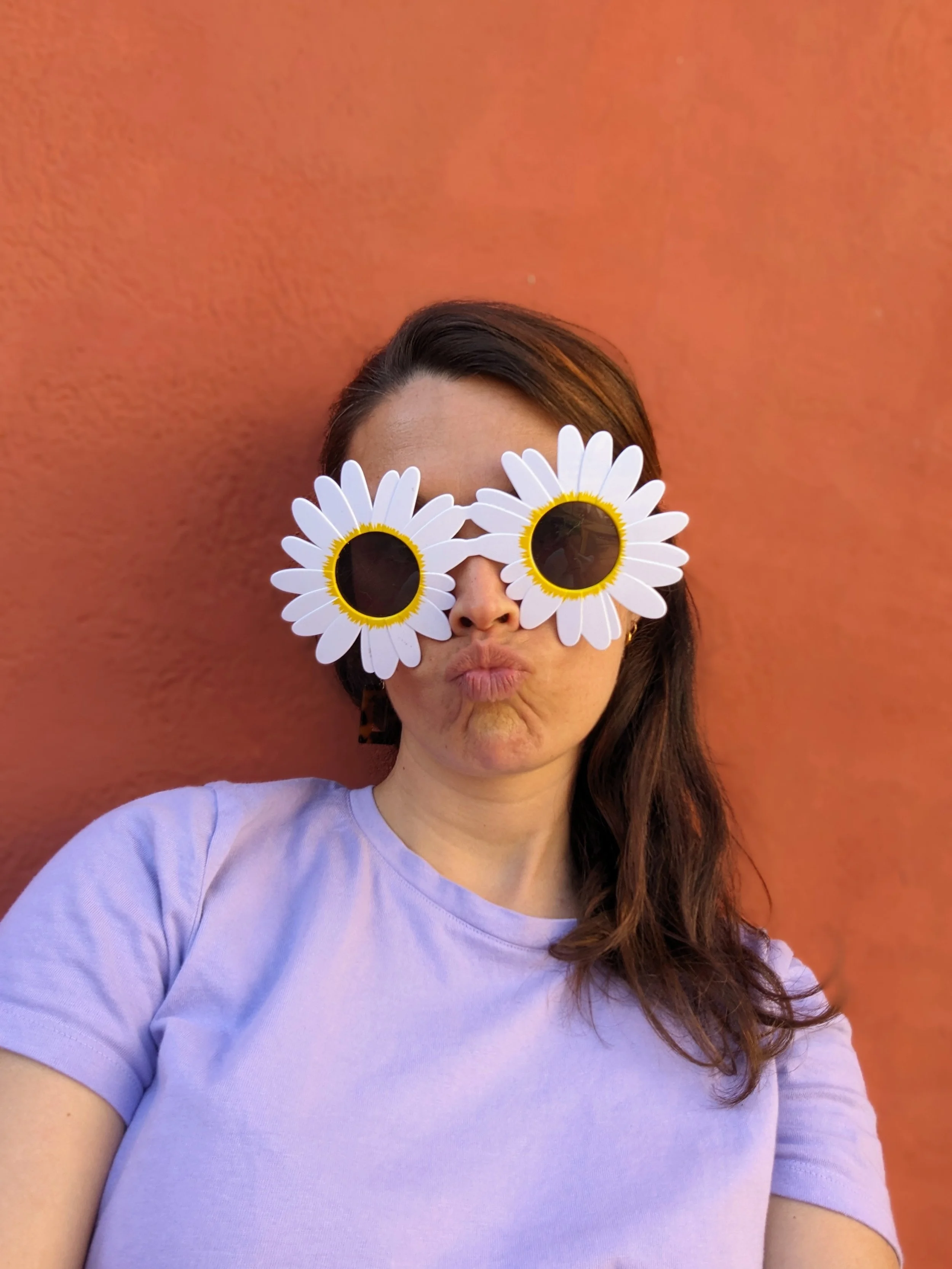 Une femme porte des lunettes en forme de marguerite, faisant un bisou, devant un mur orange.