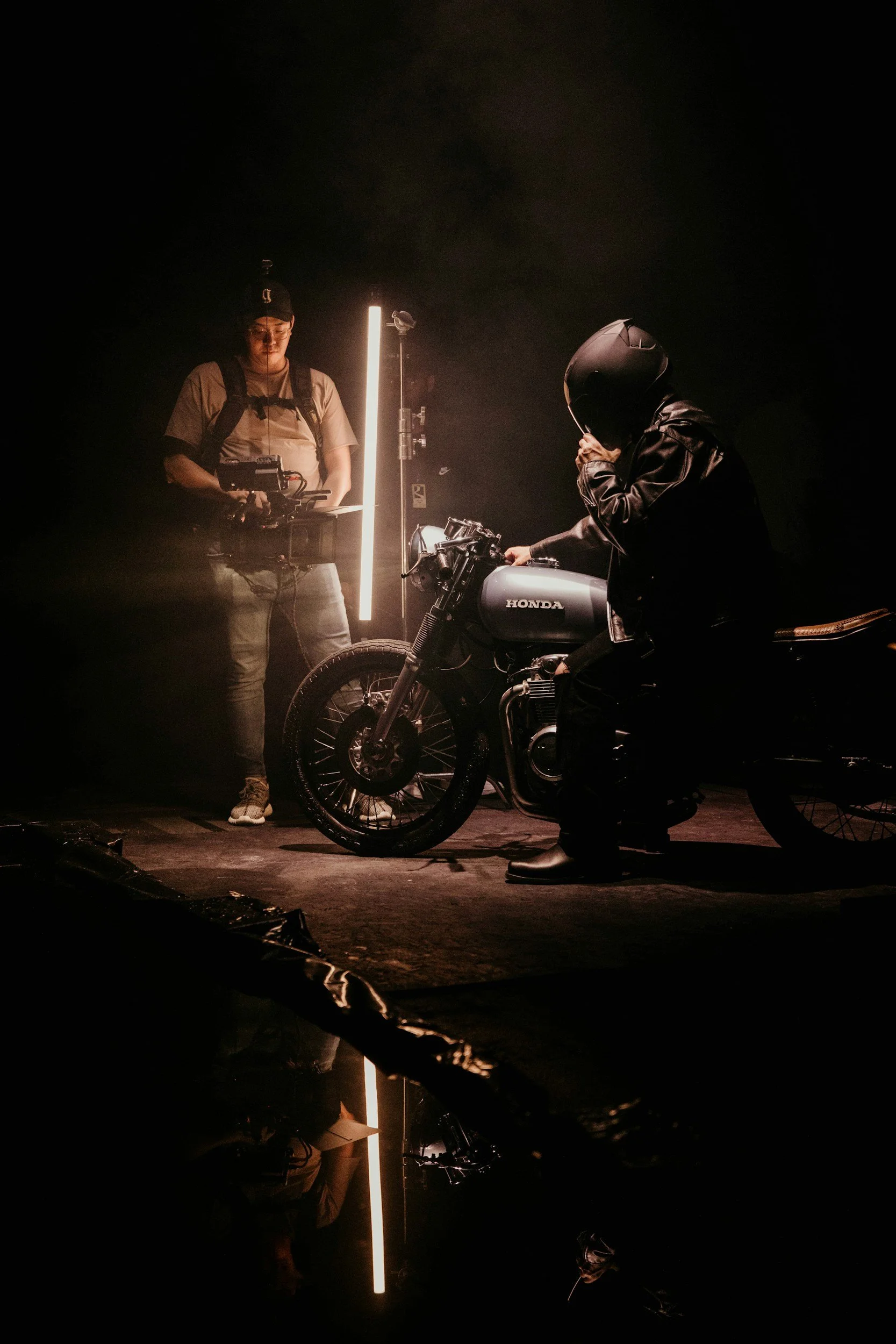 Person wearing a helmet sitting on a Honda motorcycle in a dark setting with a crew member standing nearby, holding equipment, illuminated by a vertical light.