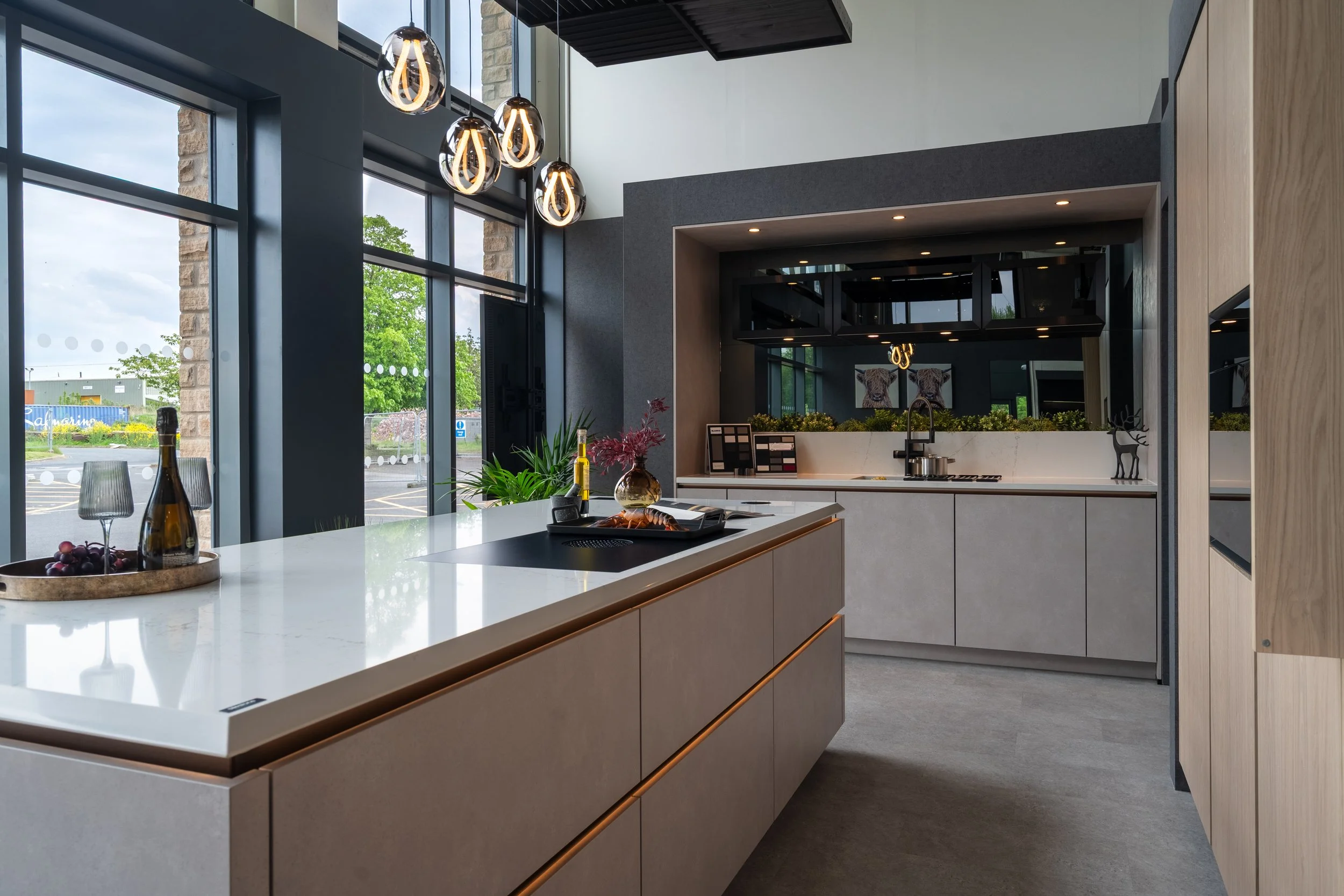 Modern kitchen with large windows, hanging pendant lights, a white island with a bottle of wine, grapes, and a tray, and a gray and wood cabinetry with decorative items, green plants, and paintings of dogs.
