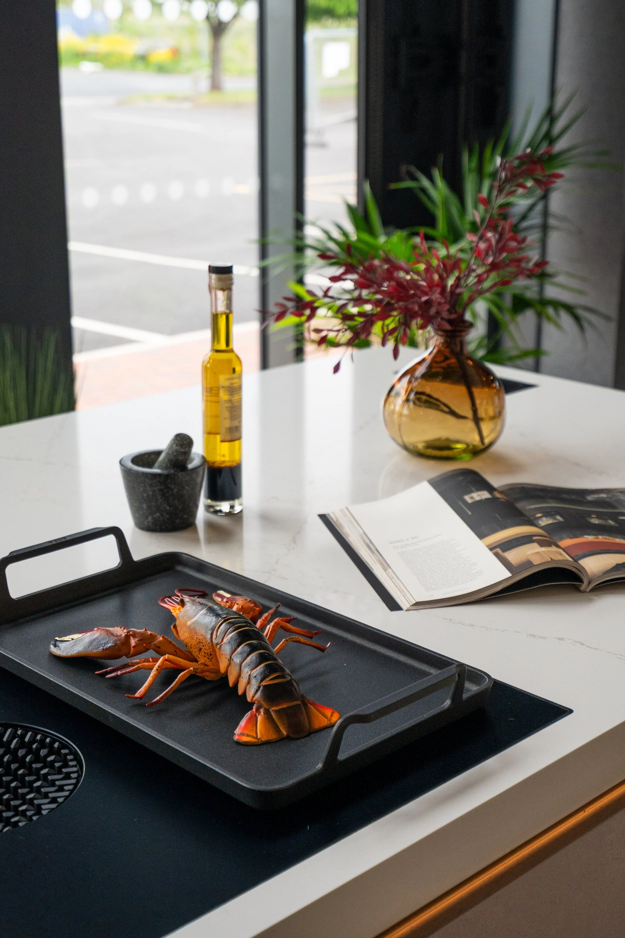 A black and red lobster and a lobster tail on a black grilling tray on a white table inside a room. In the background, there is a magazine open and a vase with red and green plants on the table. Outside the window, a parking lot and some trees are visible.