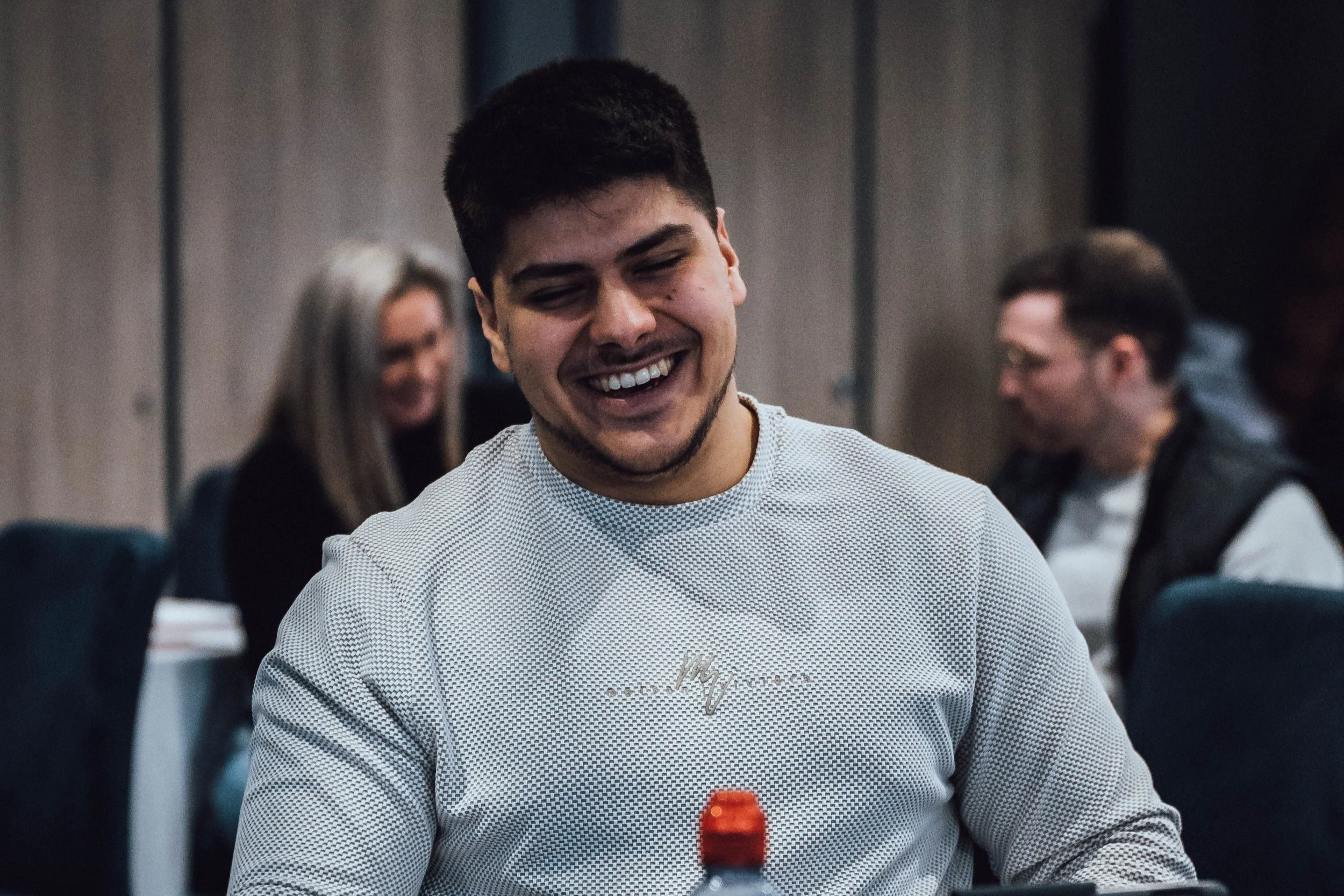 Young man with dark hair and a big smile, wearing a white checkered shirt, sitting at a table with a red bottle in front, in a room with blurred people in the background.