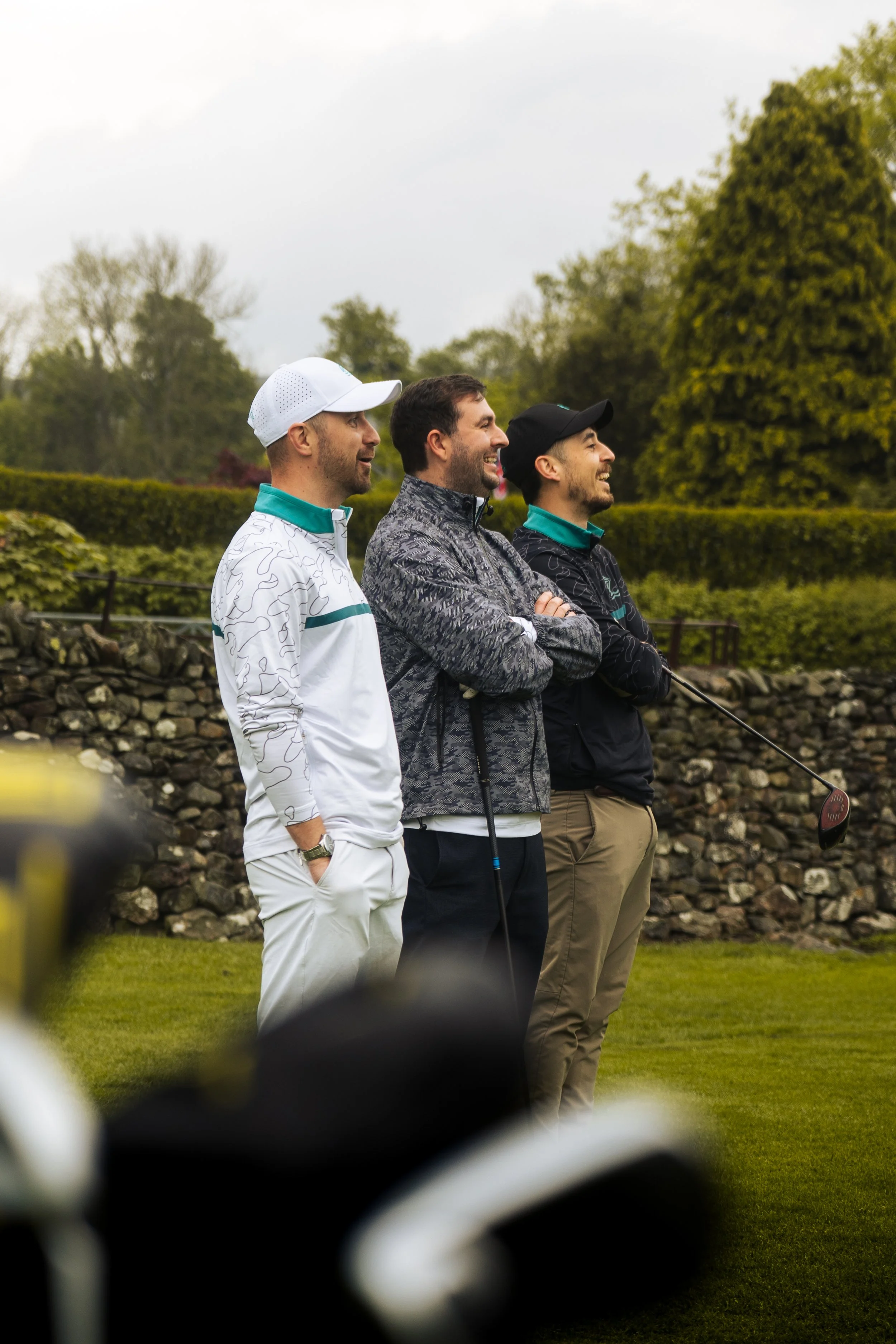 Three men standing outdoors on a golf course, watching something in the distance, with trees and a stone wall in the background.