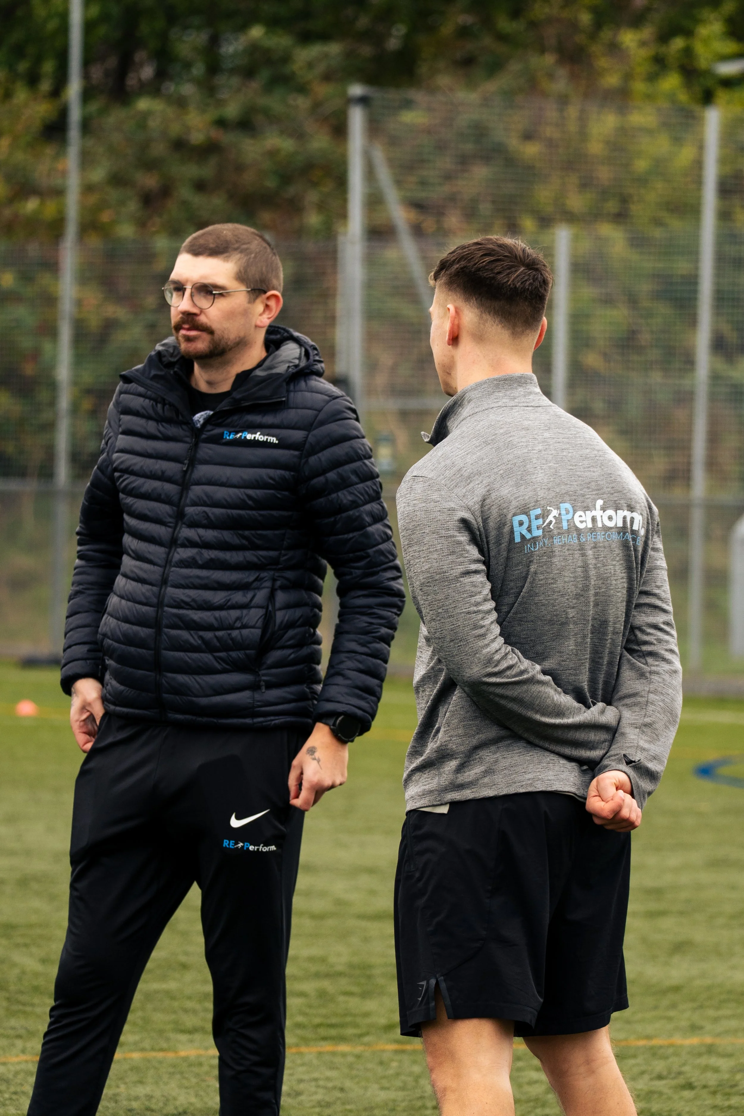 Two men standing on a grassy sports field, engaged in conversation, with a chain-link fence and trees in the background. They are wearing athletic clothing with the words "RE Perform" on their jackets and shirts.