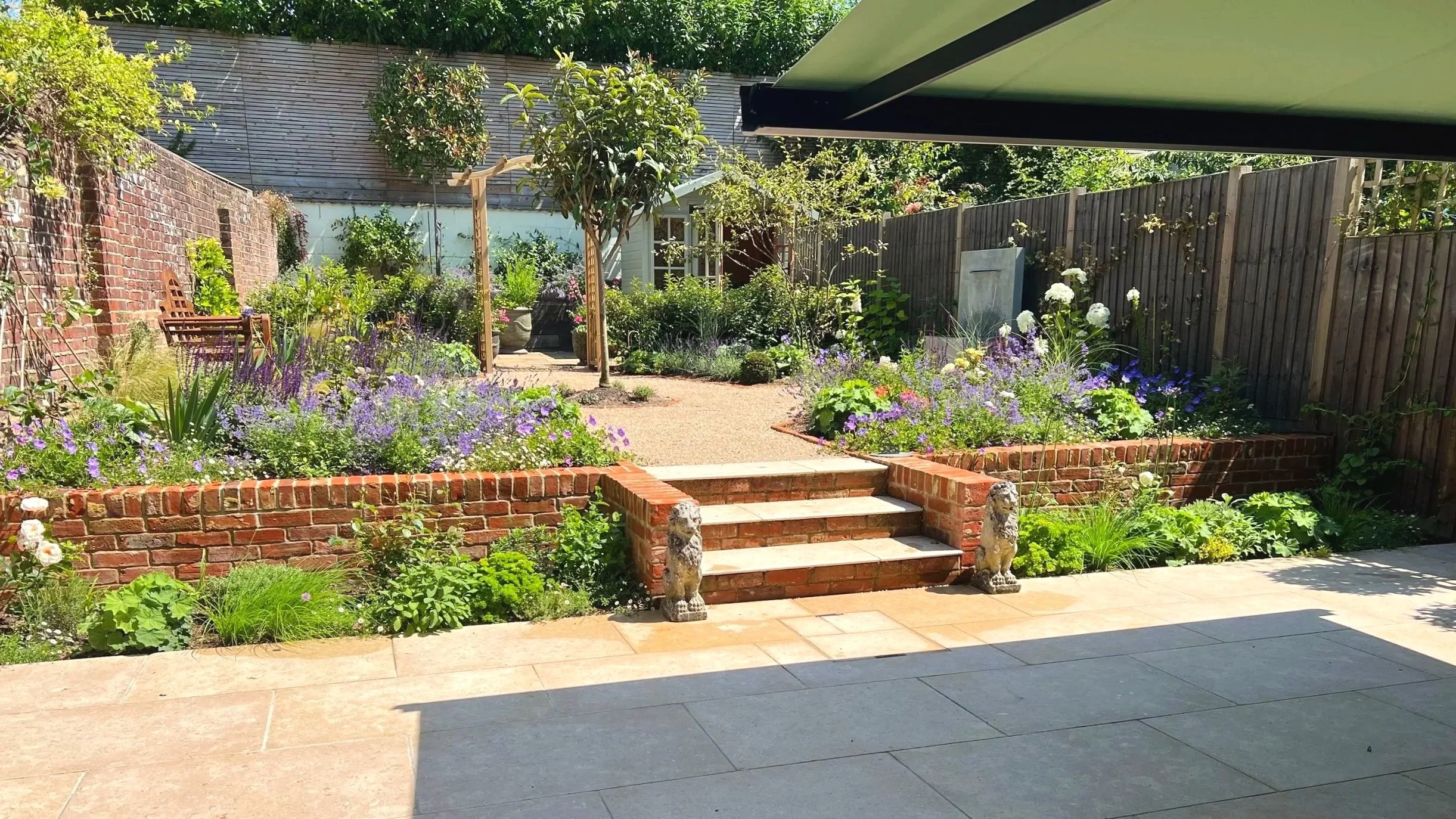 A cottage-style courtyard garden with abundant planting, a low retaining brick wall with three steps leading up to a gravelled path