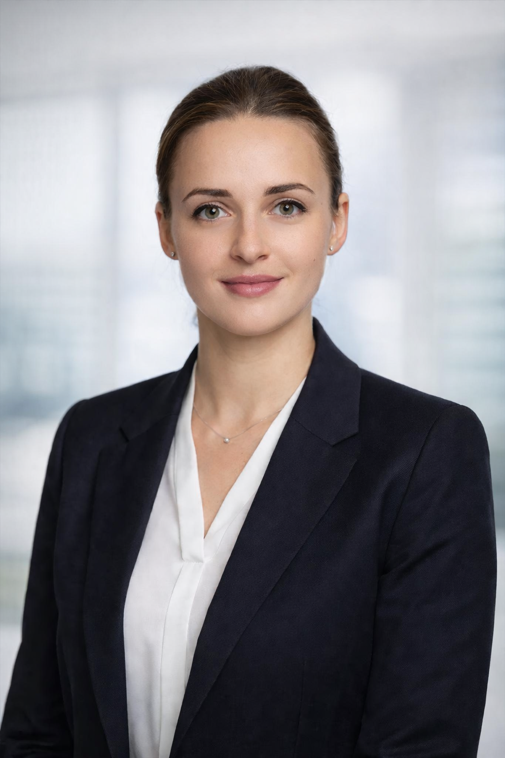A professional woman with brown hair tied back, wearing a black blazer, white blouse, and a delicate necklace, standing in front of a blurred office background.