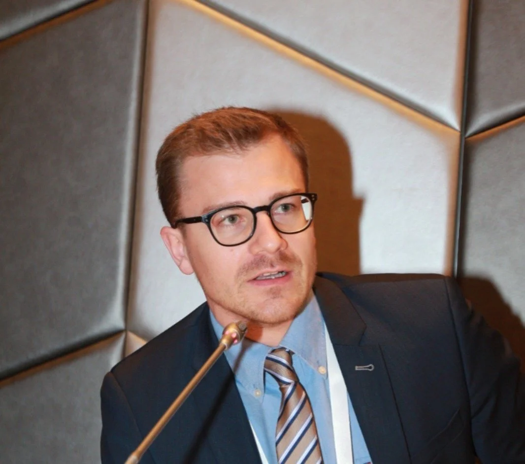 A man wearing glasses, a suit, and a striped tie speaking at a meeting or conference with a microphone in front of him.