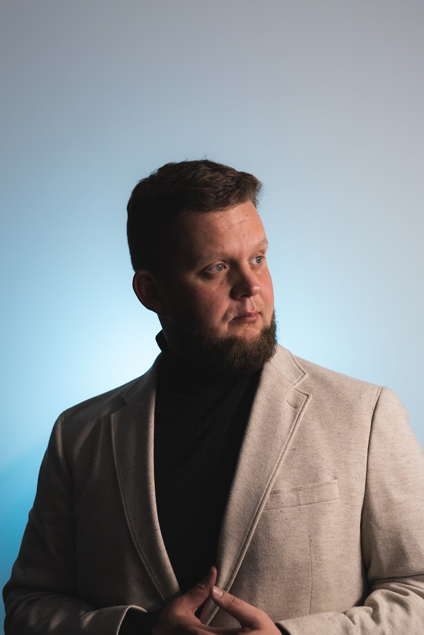 Portrait of a man with a beard wearing a beige blazer and black turtleneck, looking to the side with a neutral expression against a gradient blue background.