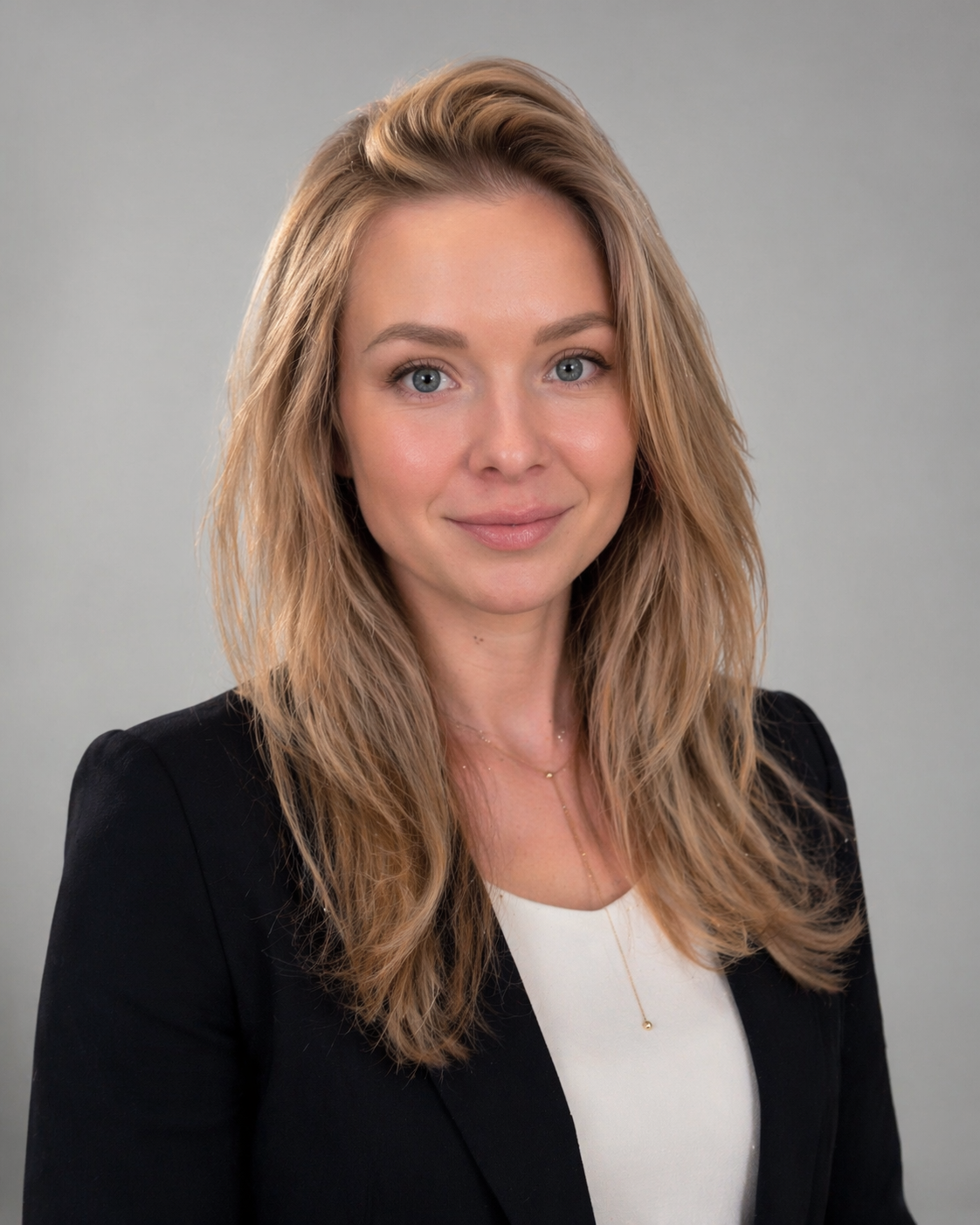 A professional woman with long blonde hair and blue eyes wearing a black blazer and white top, smiling and posing against a plain gray background.