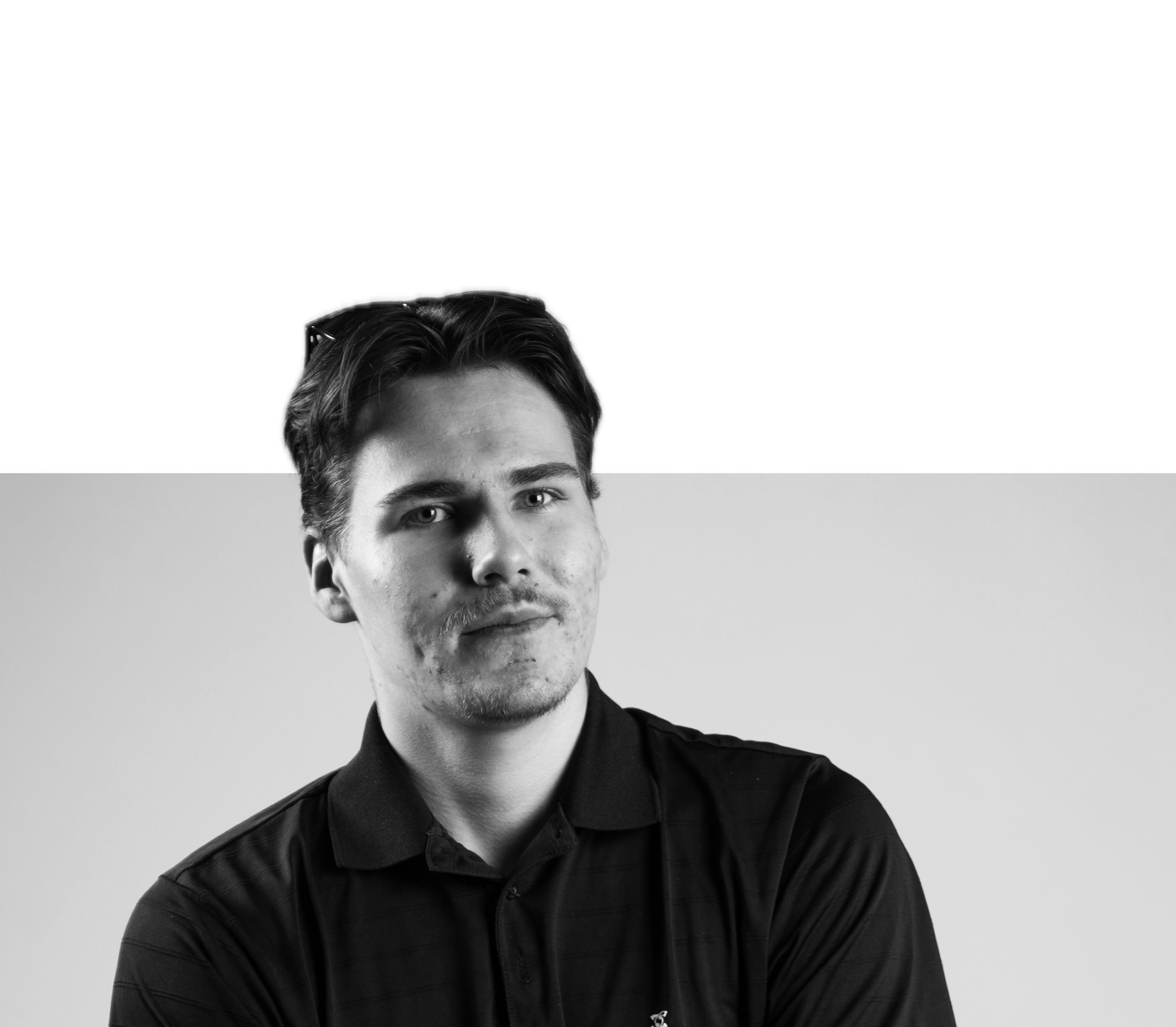 Mustavalkoinen kuva nuoresta miehestä, katse suoraan kameraan, rennossa asennossa, taustalla kaksi vaakasuoraa värilohkoa, yläosa musta ja alaosa vaaleanharmaa, hänellä tummat hiukset ja musta pikeepaita.