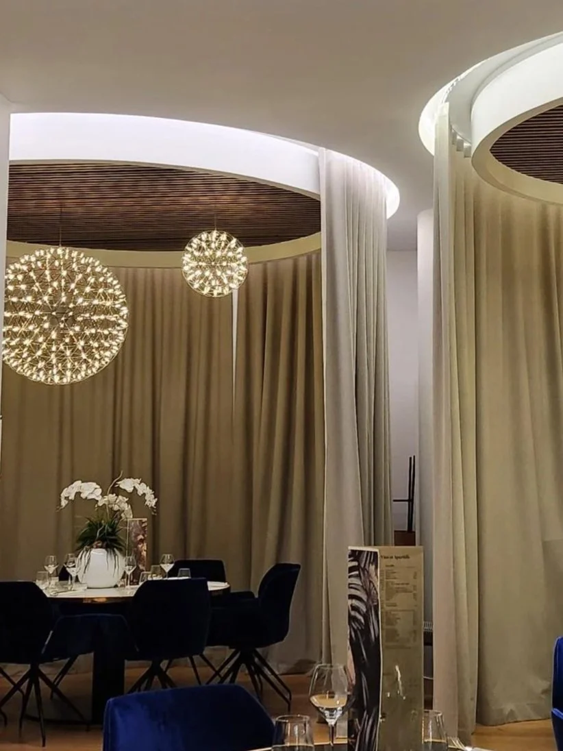 Intérieur de restaurant élégant avec des rideaux beiges, des suspensions modernes, une table ronde dressée avec des verres à vin et un centre de table floral.