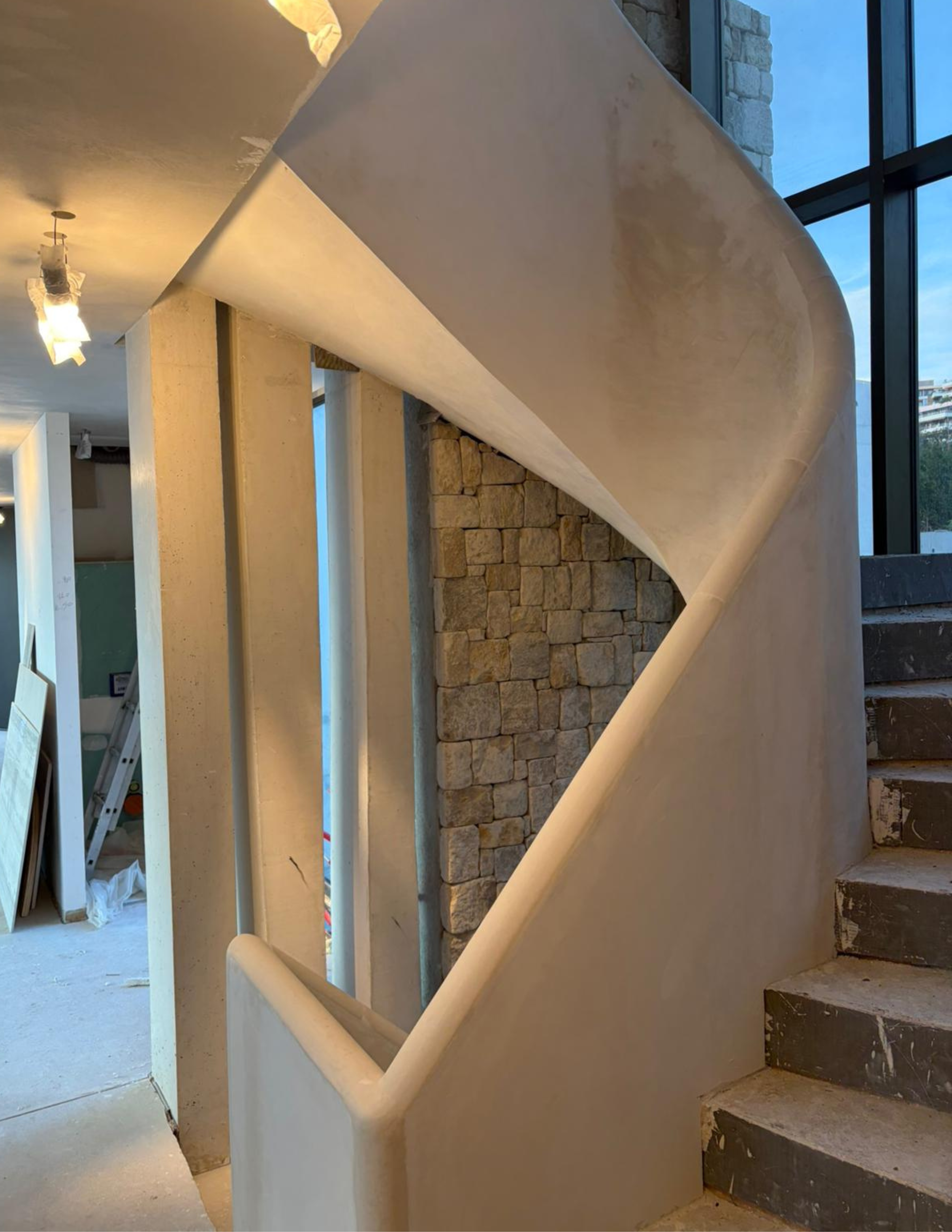 Vue intérieure d’un escalier avec une rampe blanche, courbe et lisse, un mur en pierre et de grandes baies vitrées en arrière-plan.