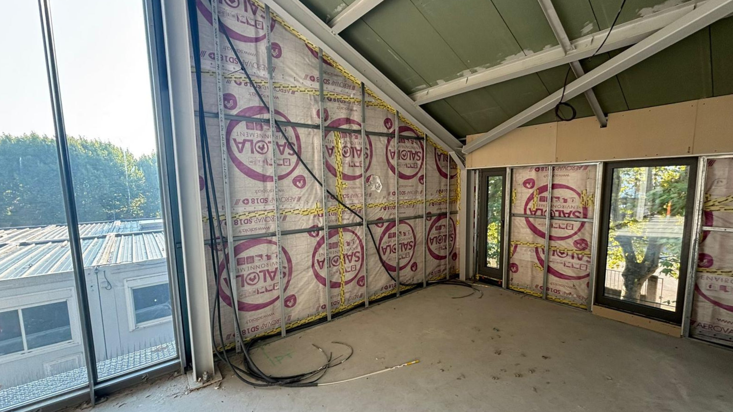 Intérieur d’un bâtiment en cours de construction, avec l’isolation et l’ossature terminées, de grandes fenêtres le long du mur et des câbles électriques apparents.