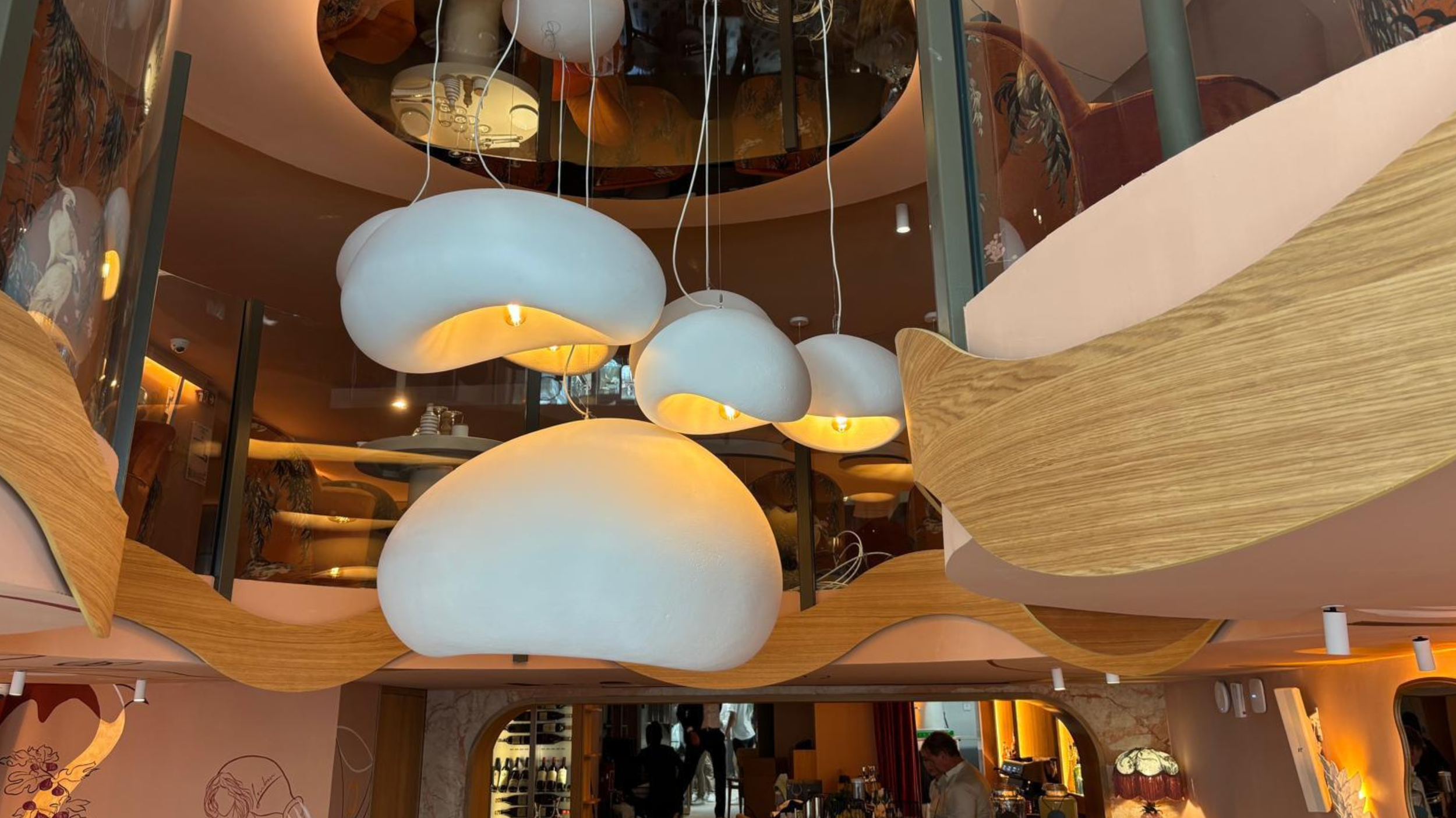 Intérieur d’un restaurant avec de grands luminaires suspendus blancs en forme de nuages, un plafond en bois au design ondulé et un éclairage ambiant chaleureux