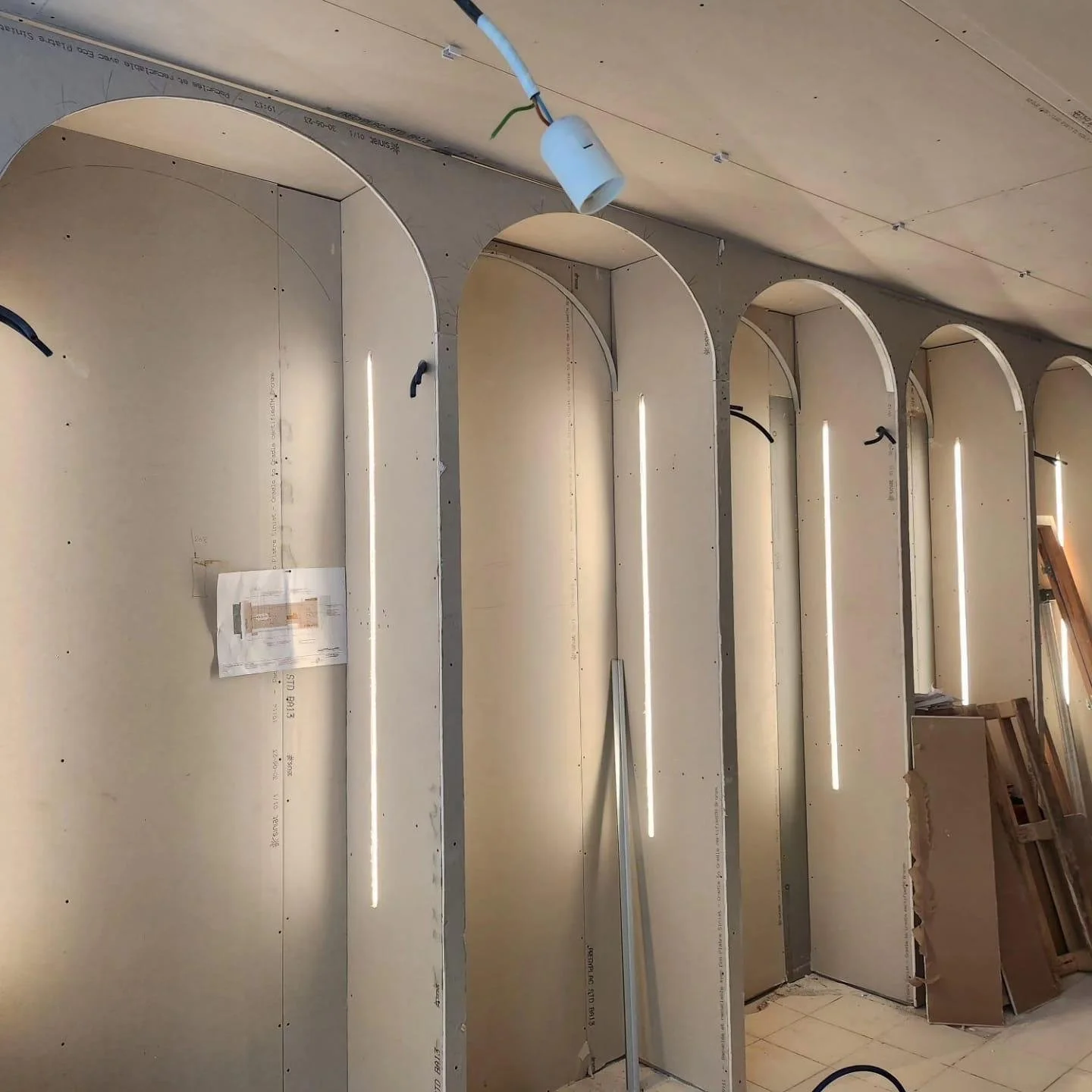 Chantier de construction avec des arches en plaques de plâtre courbées, installation de bandes LED et des matériaux de construction empilés au sol.