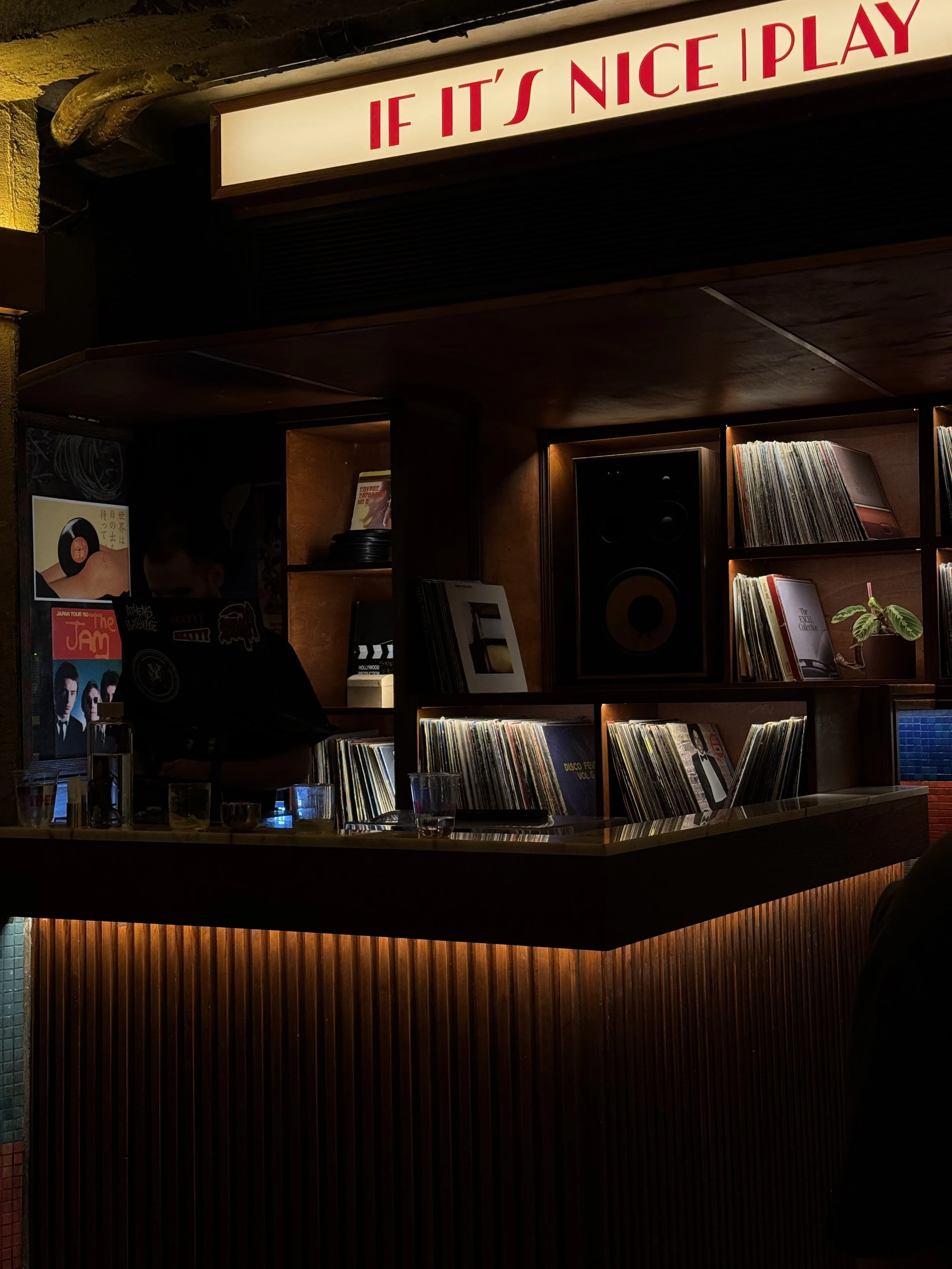Interior of a dimly lit bar or record store with a wooden counter, vinyl records on shelves, a speaker, and a sign that reads "If It's Nice I Play."