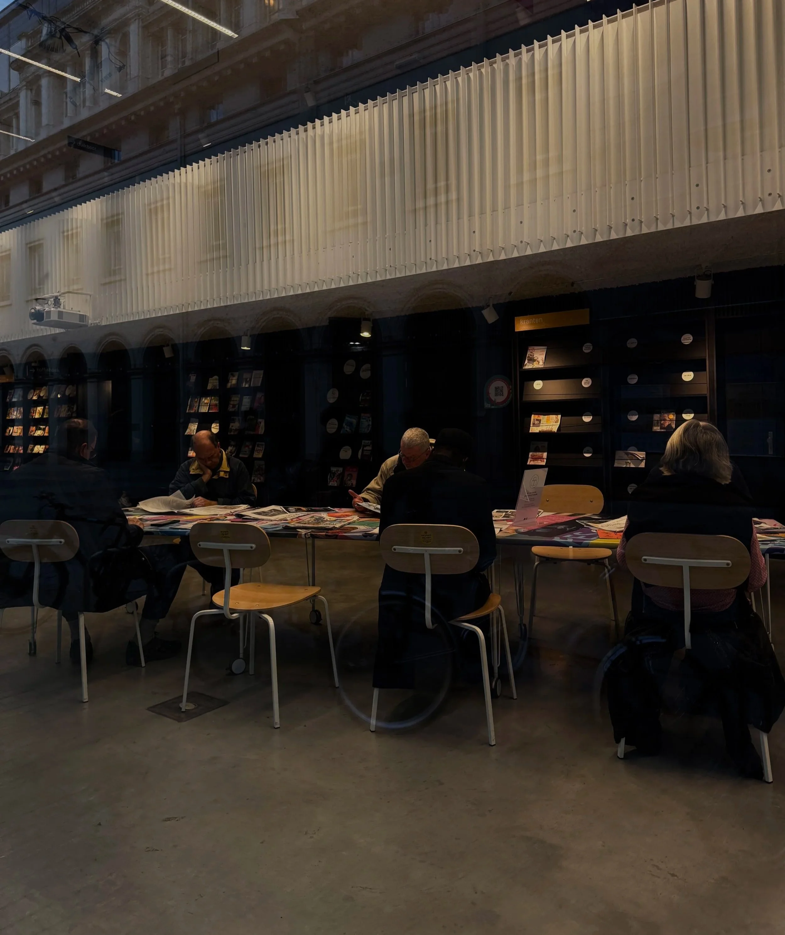 People sitting at a long table inside a bookstore, reading or browsing magazines, with shelves of magazines and books behind them, viewed from outside through a glass window.