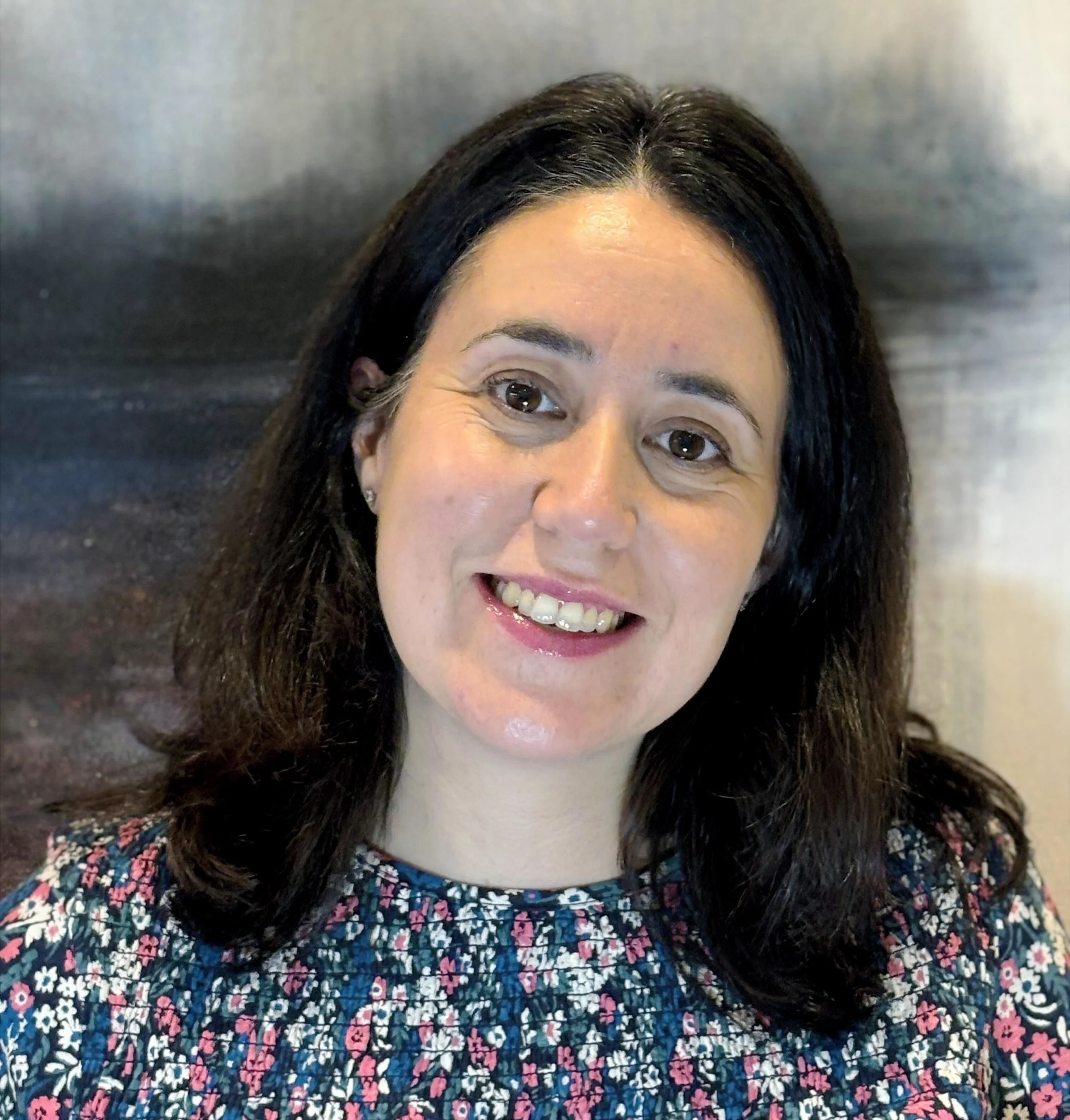 Portrait of a woman with dark brown hair, smiling, wearing a patterned top with pink, white, and blue flowers.
