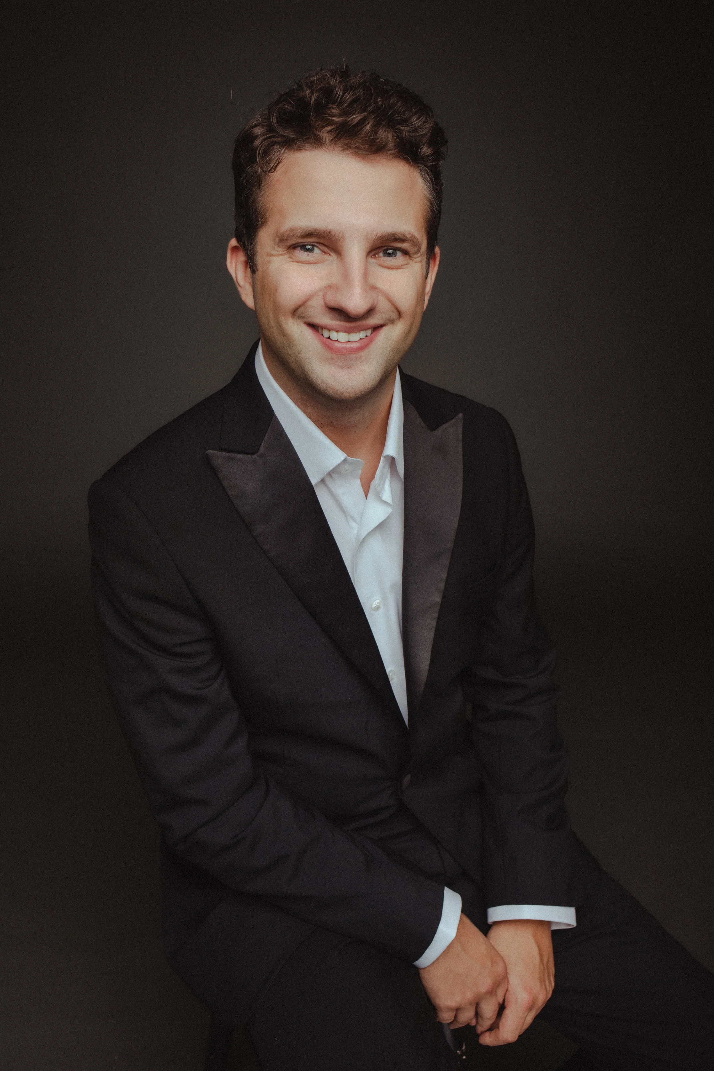 Portrait of a young man with short curly hair, wearing a black tuxedo and white shirt, smiling against a dark background.