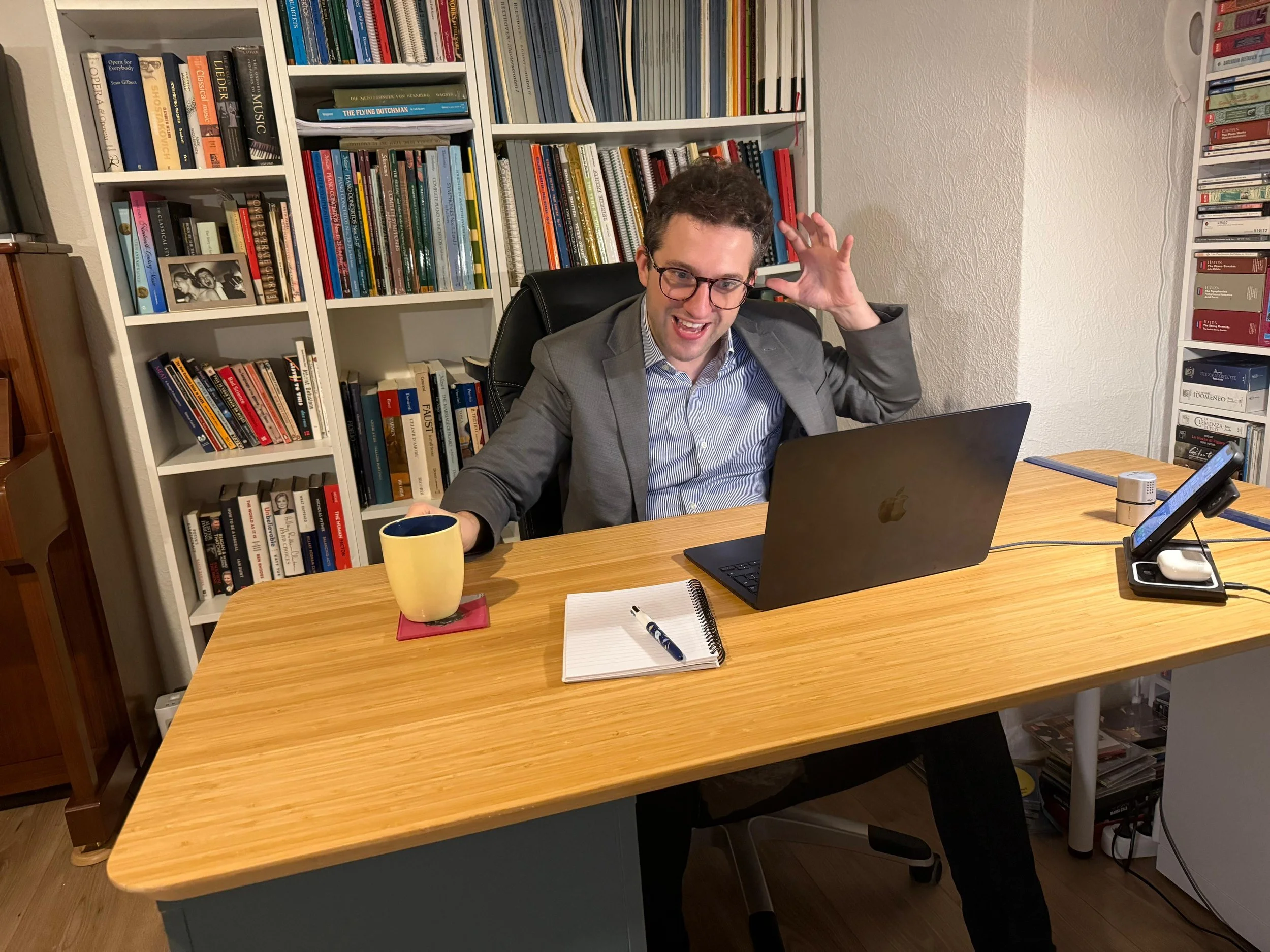 A man in a gray suit and glasses sitting at a wooden desk with a black laptop, a yellow mug, a notepad with a pen, a smartphone, and a small speaker. He is smiling and making a playful gesture near his head in a room with bookshelves filled with books.