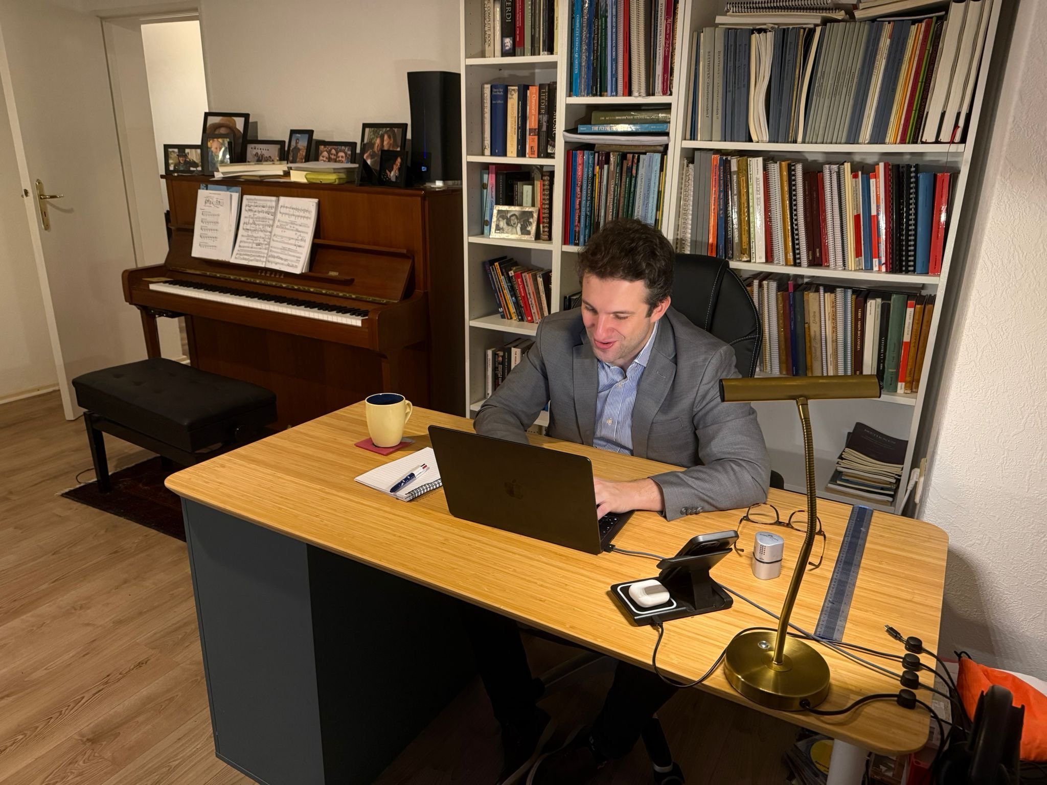 A man in a gray suit sitting at a wooden desk, working on a laptop, with a notebook, pen, and coffee mug nearby, in a home office with a bookshelf and a piano in the background.