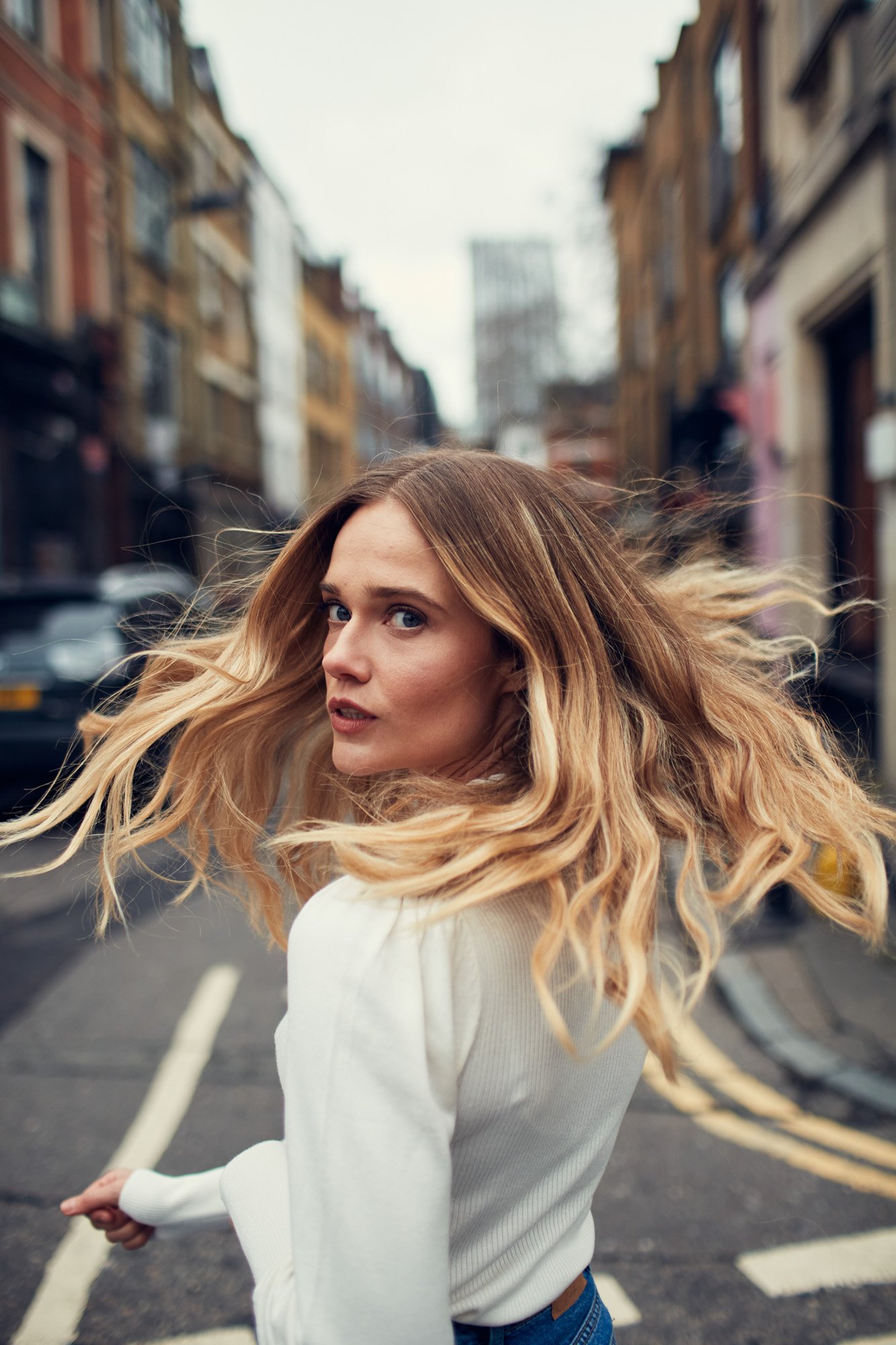 Singer Florrie, with long, wavy blonde hair and blue eyes turning her head over her shoulder on a city street in Shoreditch London, with buildings and cars in the background.