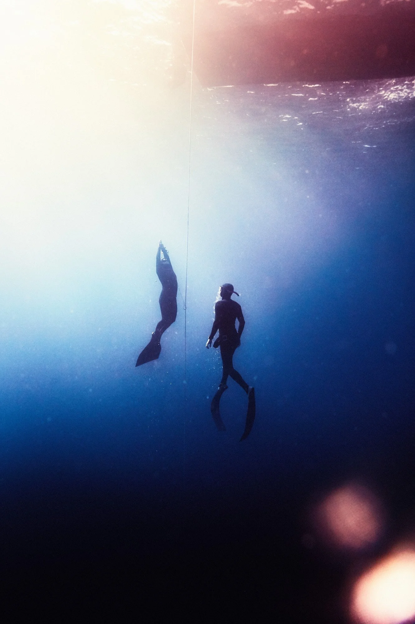 Two scuba divers underwater, one hanging from a rope and the other swimming nearby, with sunlight filtering through the water.