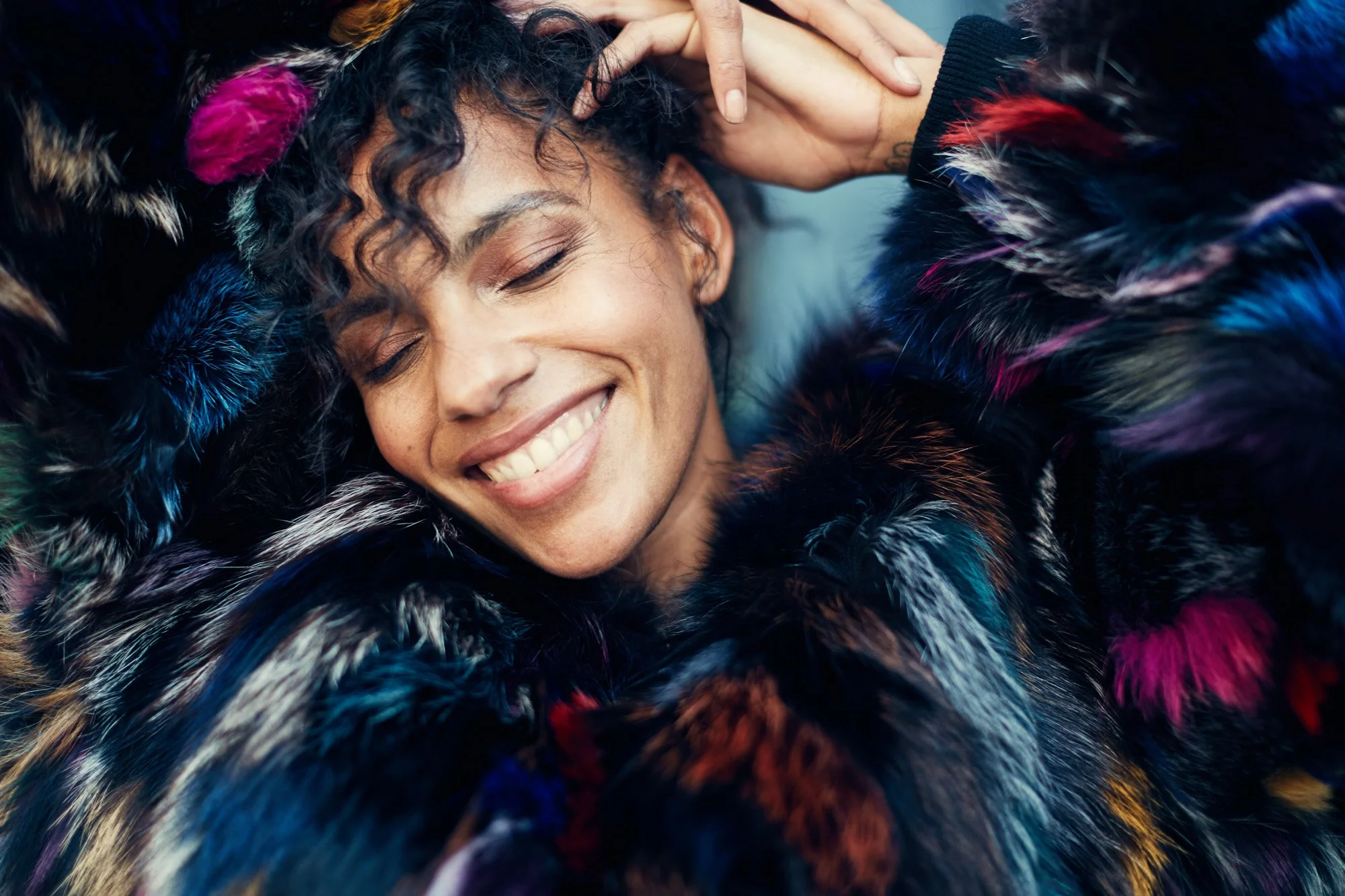 A young woman with curly hair smiling and resting her head on a colorful fur coat.