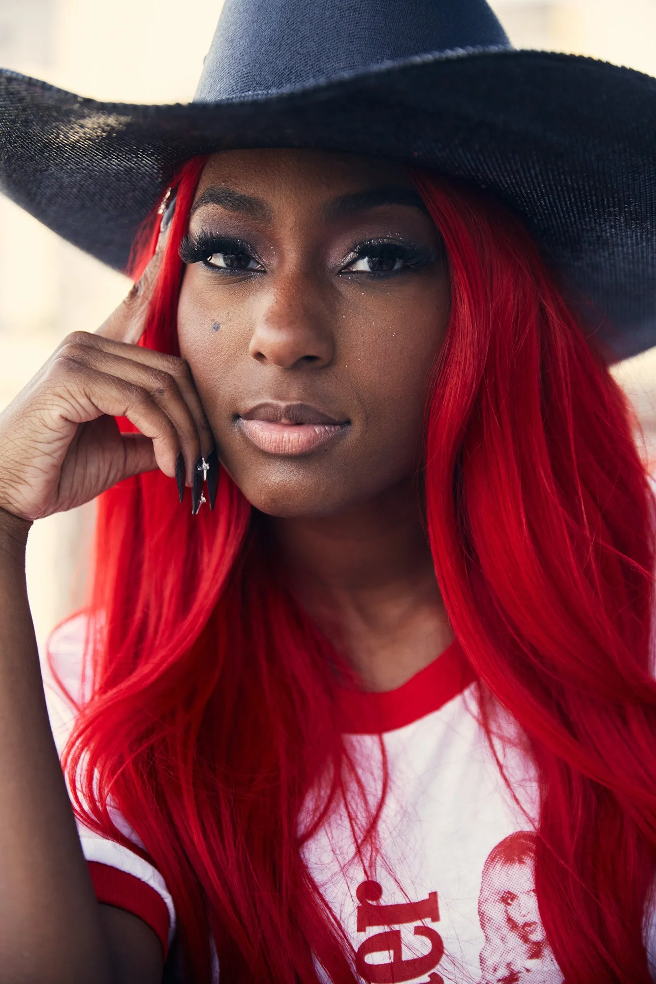 Close-up portrait of singer Reyna Roberts with long red hair wearing a black wide-brimmed hat and a white and red T-shirt featuring a vintage image of a woman and text. She is resting her face on her hand, with makeup and long false eyelashes.